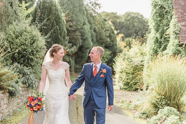 Bride and groom walking together, smiling hand in hand down a lane lined with green trees and borders