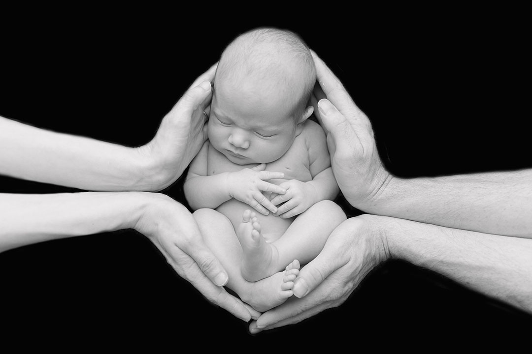 Black and white photo on a black background, baby is posed and curled up womb like iand held in their parents hands from each side