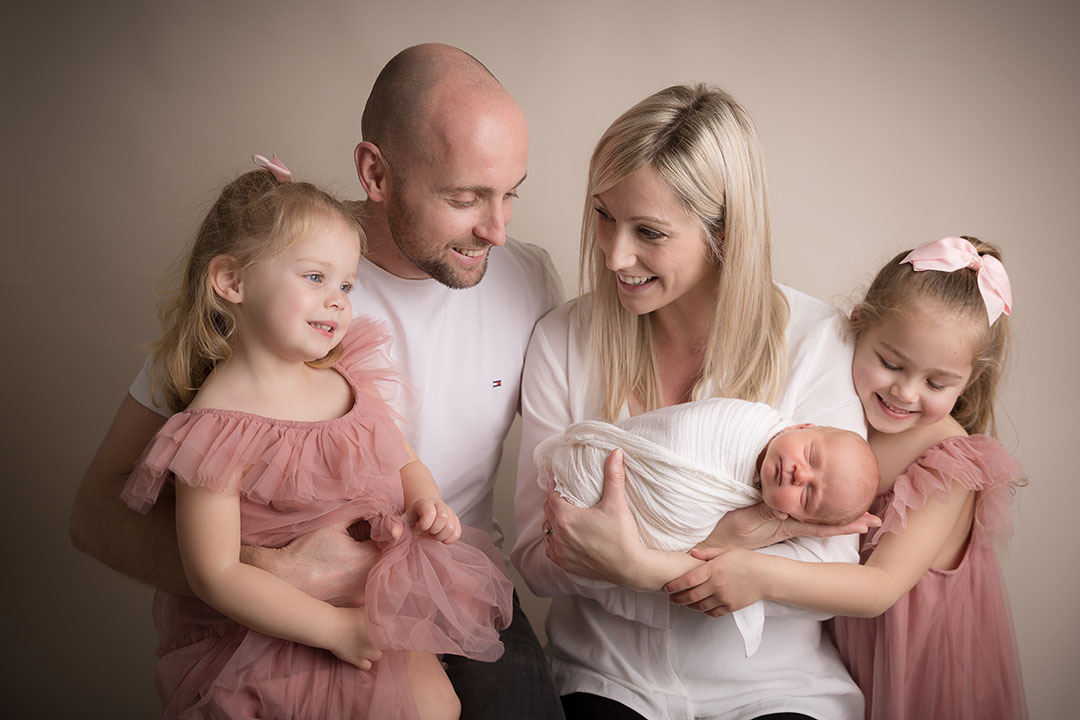 Family of 5, mum, dad, two older siblings wearing pink dresses posed with their new baby brother wrapped in a white wrap against a neutral backdrop in a photography studio at hayley morris photography 