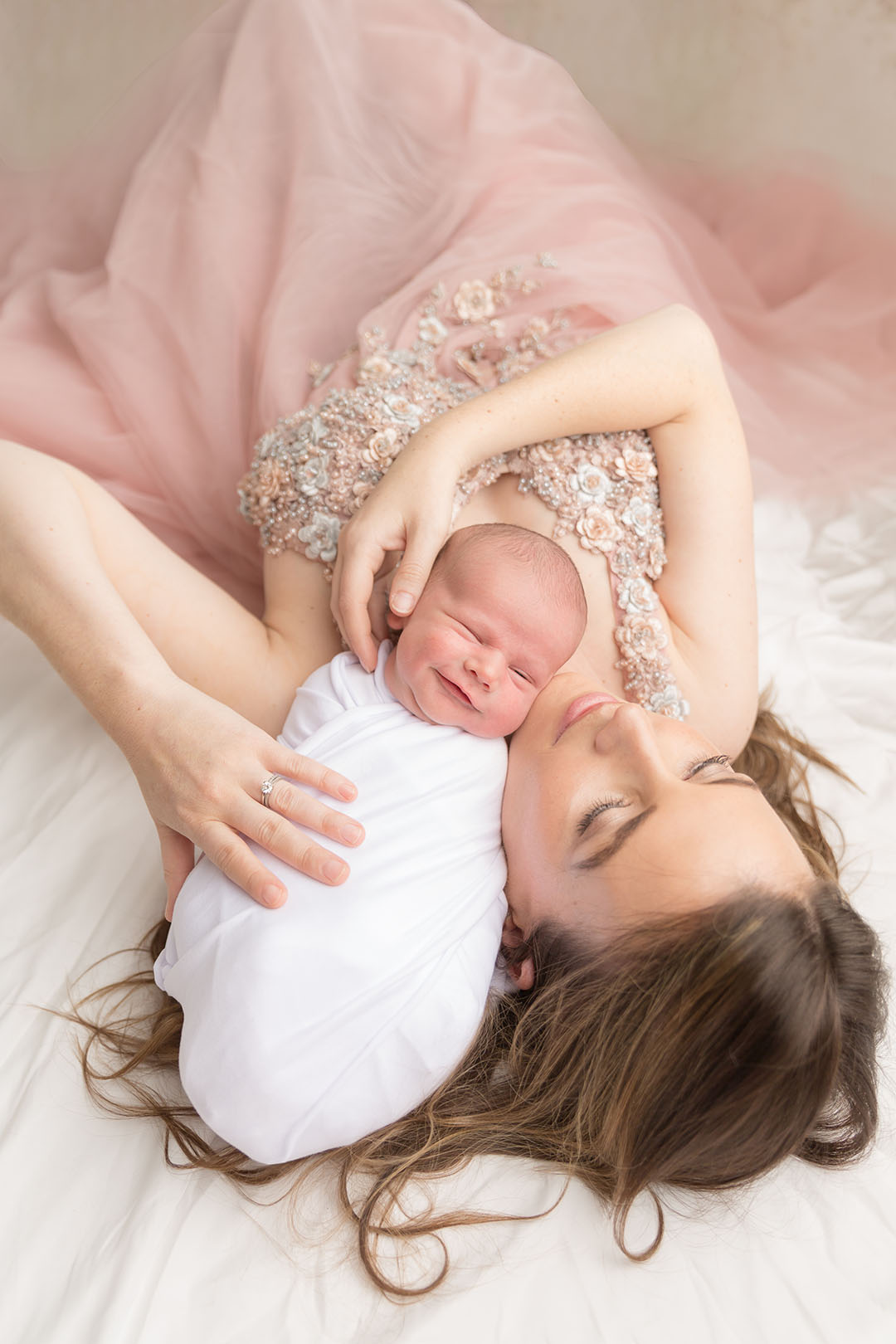 New mum wearing a gown with beads and floral detail on the bodice with tulle skirt. She's lying posed with her new baby wrapped in a white wrap. baby is smiling softly 