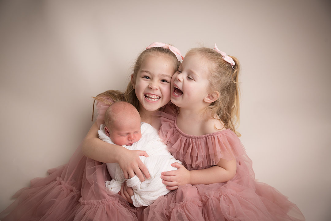 Baby wrapped in a white wrap at their newborn session. Photograph is taken against a neutral backdrop with two young girls (the baby's siblings) holding him. They are smiling/laughing and dressed in pink tulle dresses