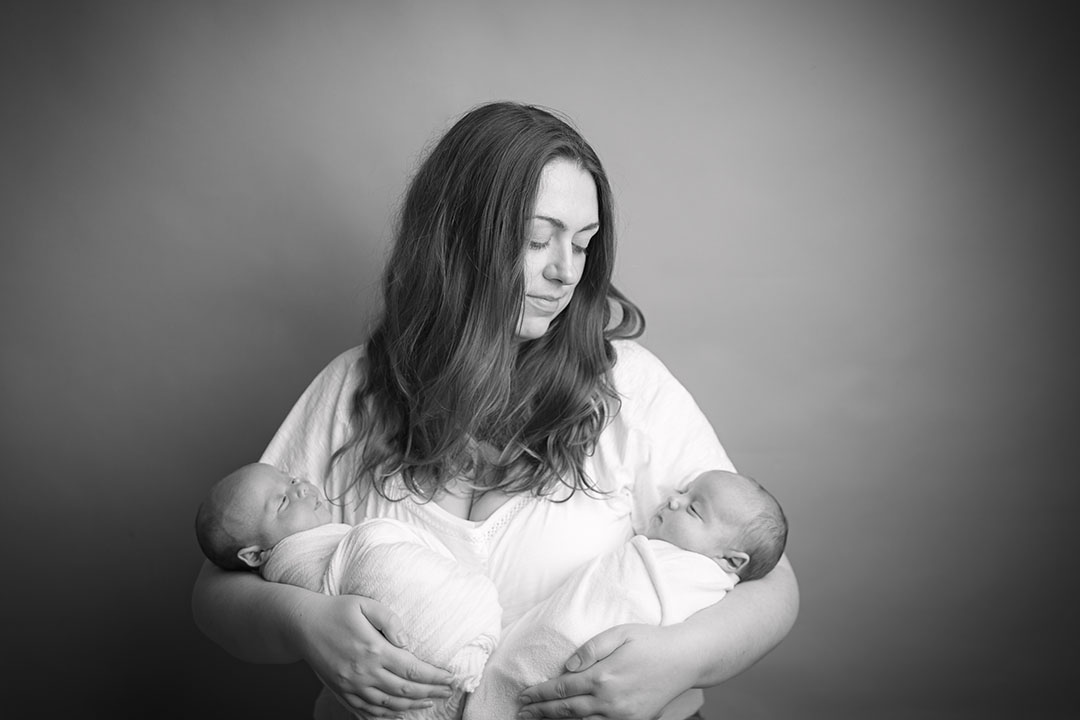 New mum holding her identical twin babies having her portrait taken. Babies are wrapped in cream/white fabric. mum is wearing a white top