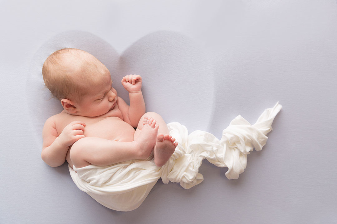 baby lay partially wrapped and posed in a heart shape over a lilac/grey fabric 