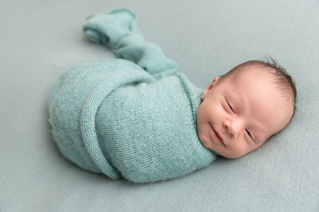 Baby wrapped in a teal fabric lay on a lighter teal fabric backdrop on a posing bag. Newborn baby is smiling 