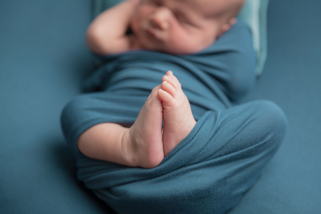 close up photo of a newborn baby's feet. baby is wrapped in a deep teal wrap and lay on a coordinating posing fabric on a posing bag