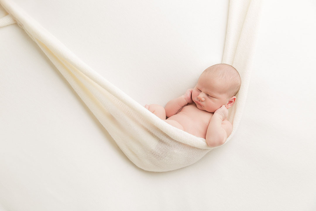Cream backdrop on a posing bag with a baby lay posed and curled sleeping in a hammock type pose with a matching cream wrap