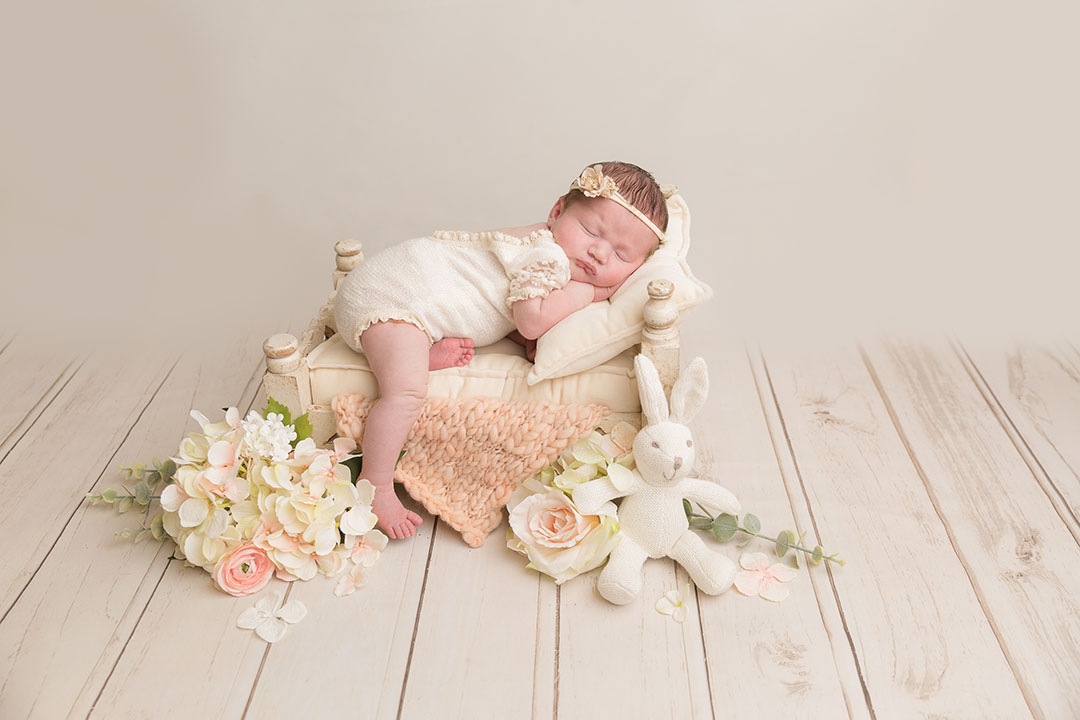 Newborn studio session on cream and wooden backdrop. Baby is wearing a vintage cream romper posed on a cream bed with headband and  her leg gently hanging over the edge. Flowers and white bunny sat at the base on the bed on the floor