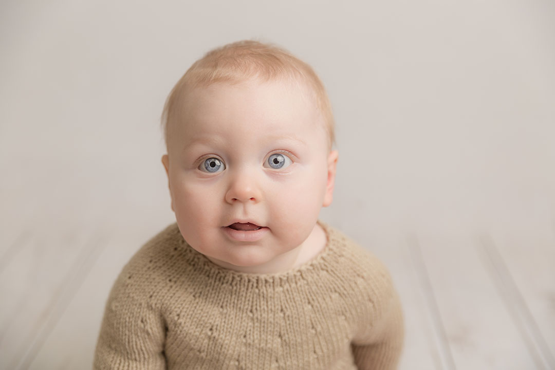 Sitter aged baby 6 7 8 9 10 months old sat being photographed wearing a knitted neutral romper against a neutral backdrop. Baby has striking blue eyes 