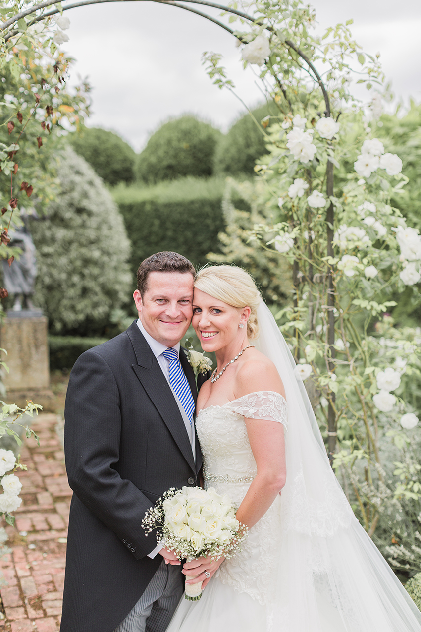 Bride and groom posed with their heads together facing the camera and hands joined in the white garden at birtsmorton court smiling in the moment