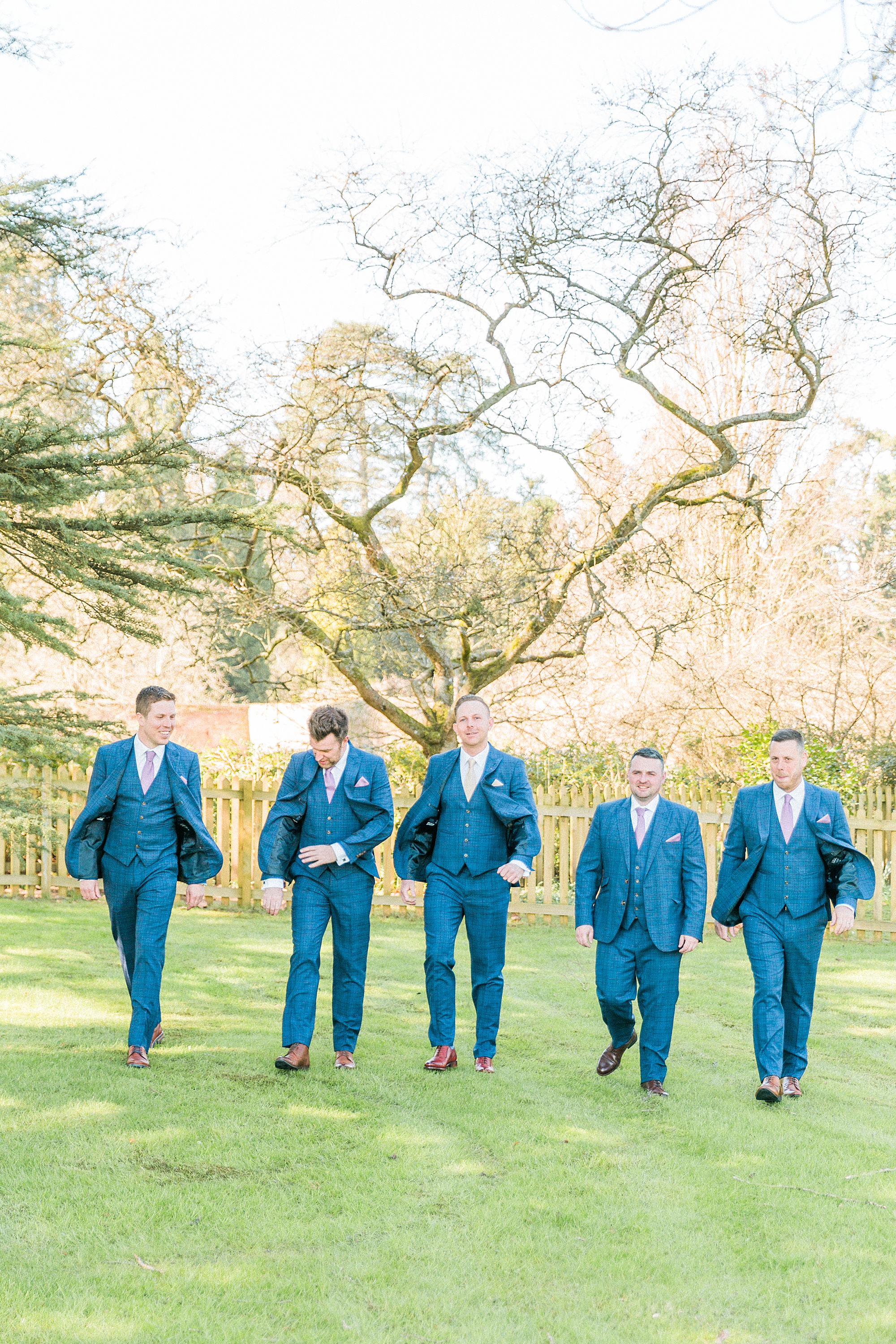 Photograph of a groom and his groomsmen dressed in suits/wedding attire walking together across a lawned area with trees talking with each other as they walk
