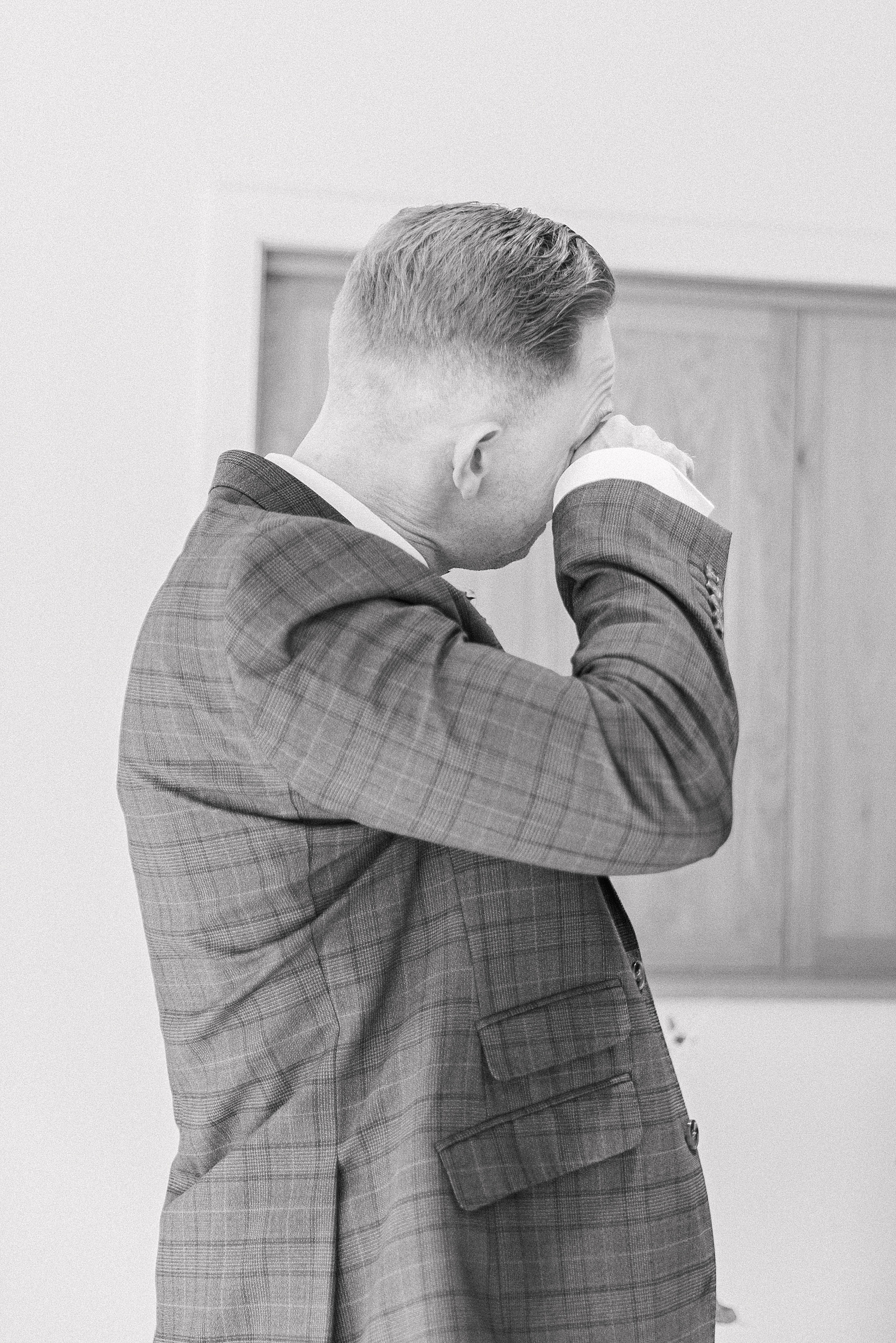 black and white photo of a groom very teary with his hand to his eyes drying a tear watching the bride as she walks down the aisle to be married (the bride isn't in the photo) 