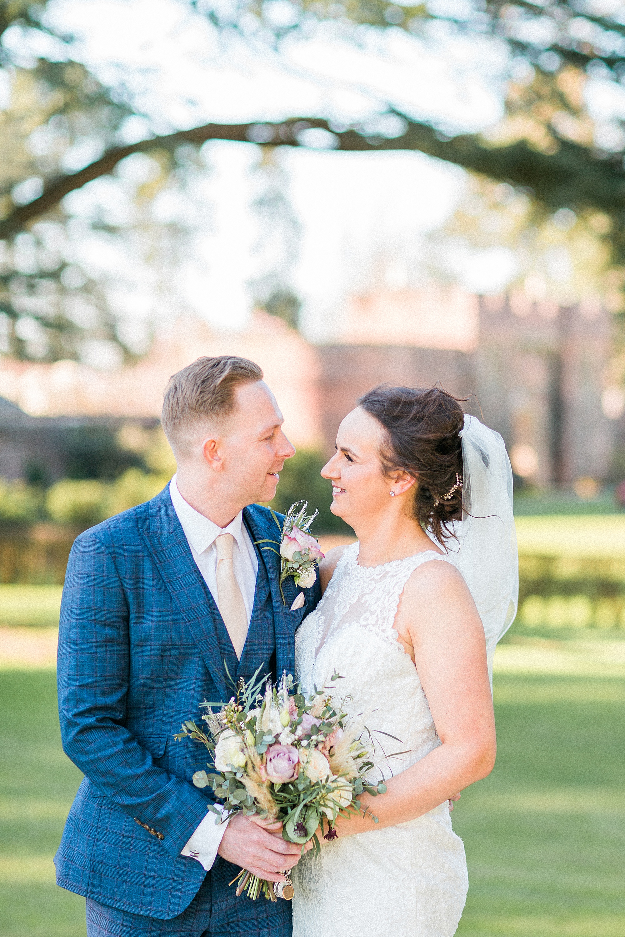 PHOTO of a bride and groom facing in towards each other smiling together on a lawned area, they've joined hands holding the bride's bouquet 