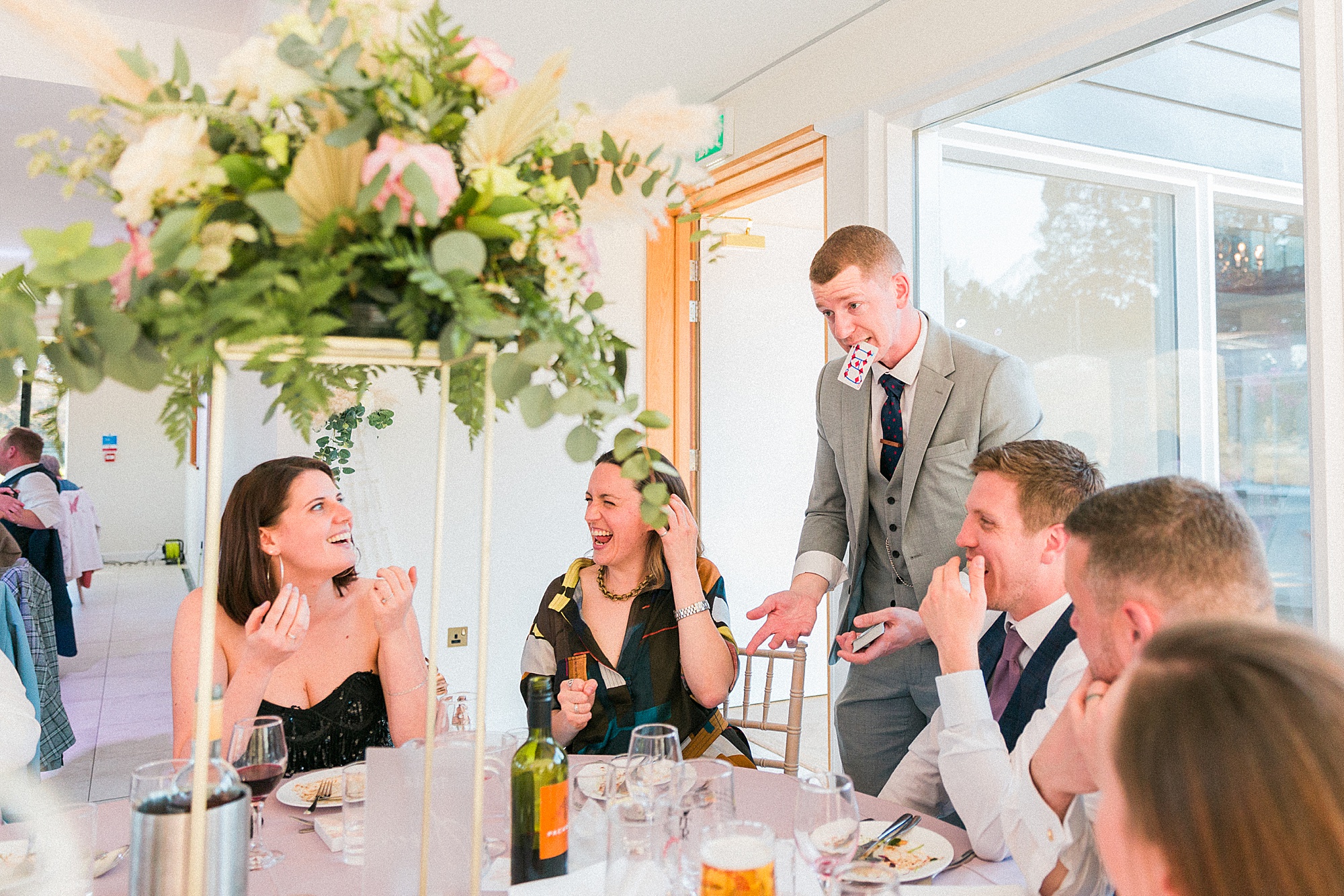 photo shows a magician with a card in his mouth and the guests sat at a table and their reactions to his trick