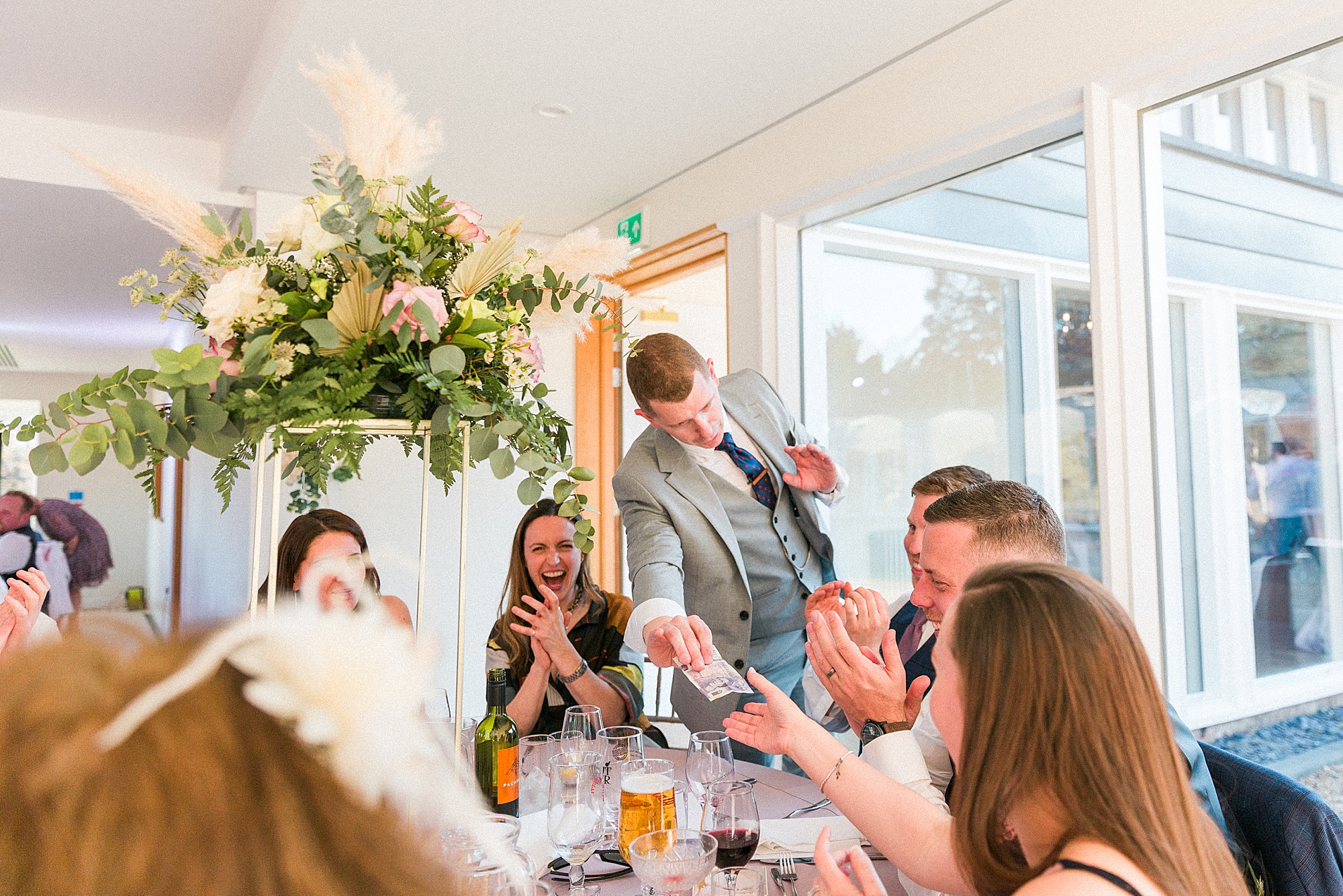 photo shows guests sat at a table in a wedding reception with a magician presenting his show to them and their reactions 