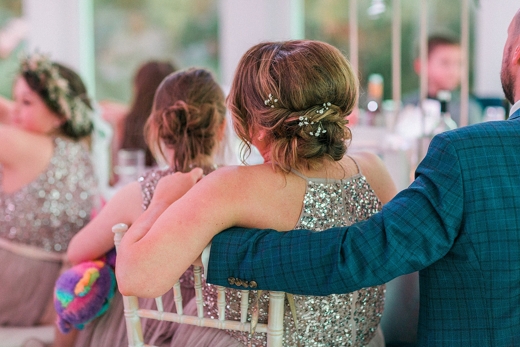 photo shows the back of wedding guests sat during the speeches. A member of the bridal party is sat next to his wife who is a bridesmaid with his arm around her 