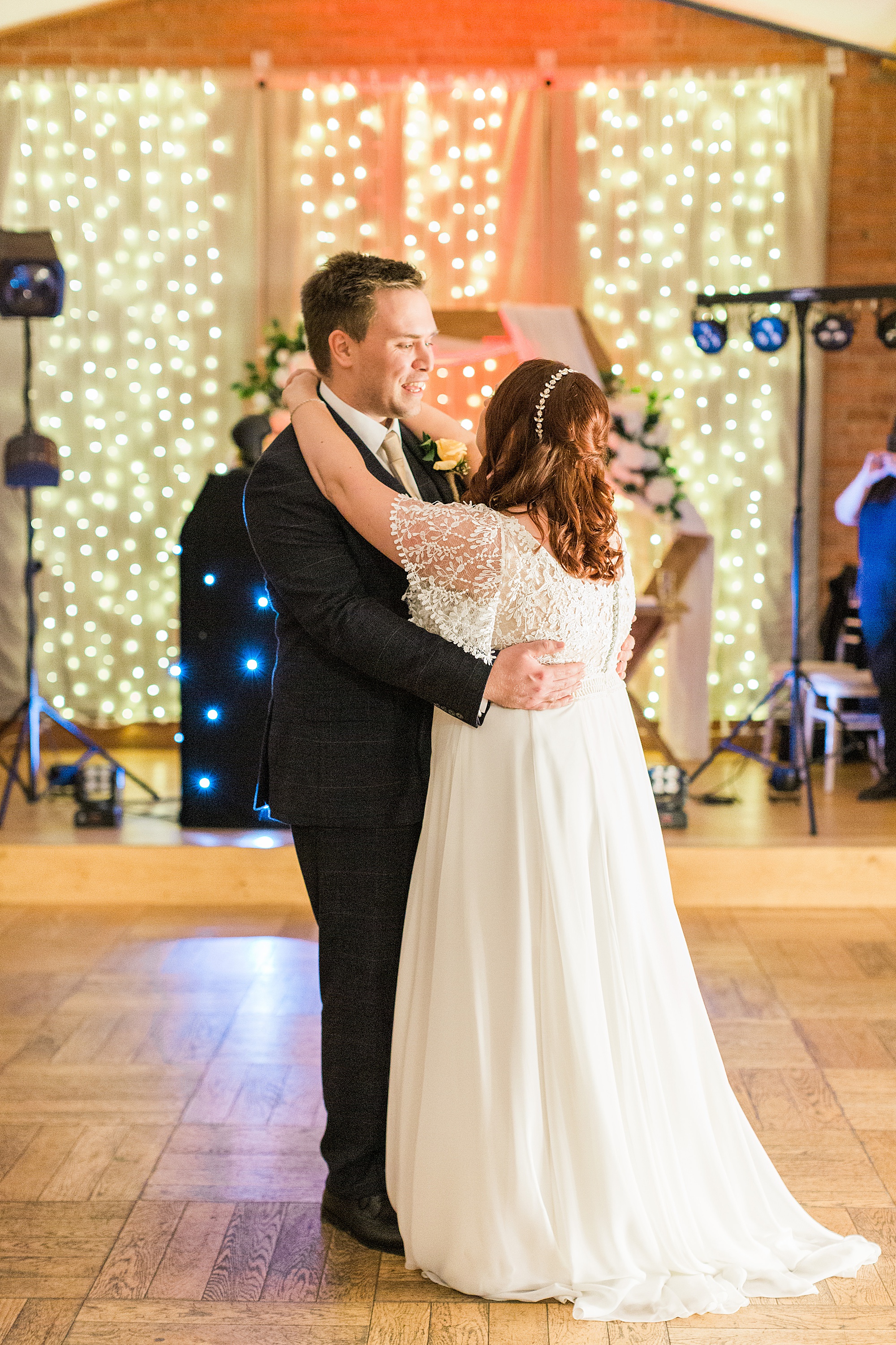 Photo of a bride and groom dancing their first dance at their wedding in at cider mill barns near stratford. Couple are backlit by fairy lights and closely embraced