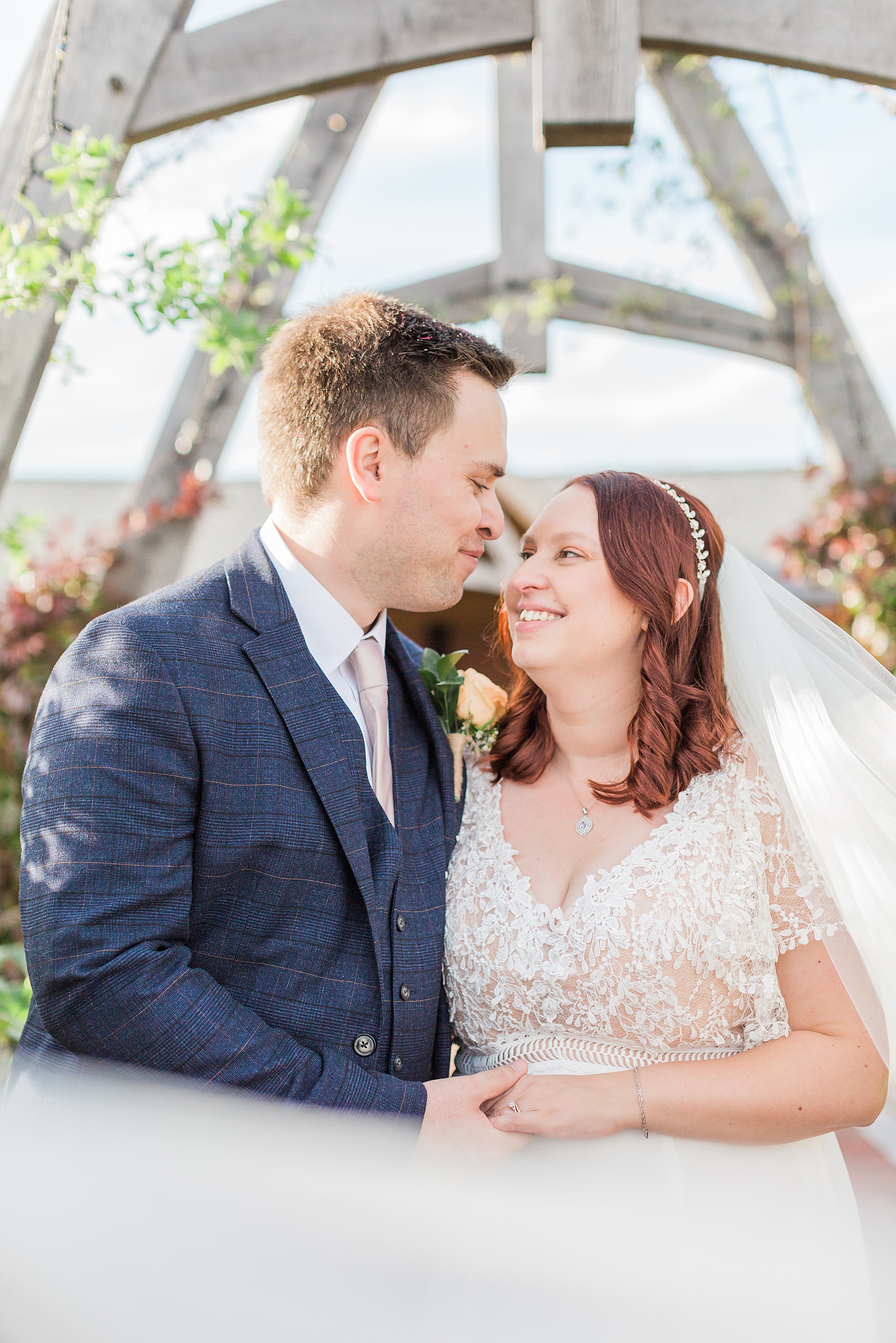 photo shows a bride and groom embraced under a wooden arch at cider mill barns. the bride's veil is in the foreground bring swept towards the camera