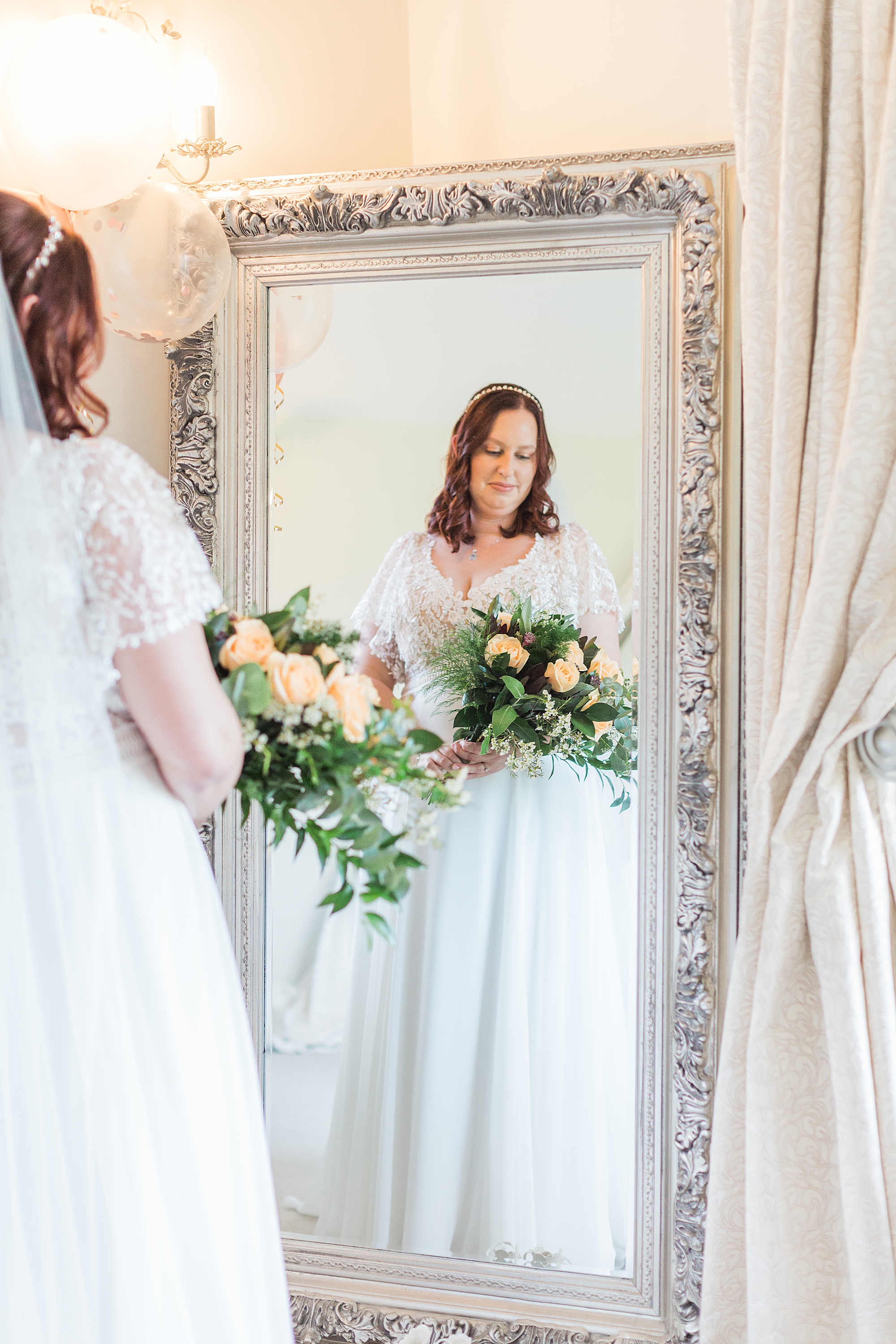 Photo shows a bride in her wedding dress holding her flowers gazing into the mirror before her ceremony 