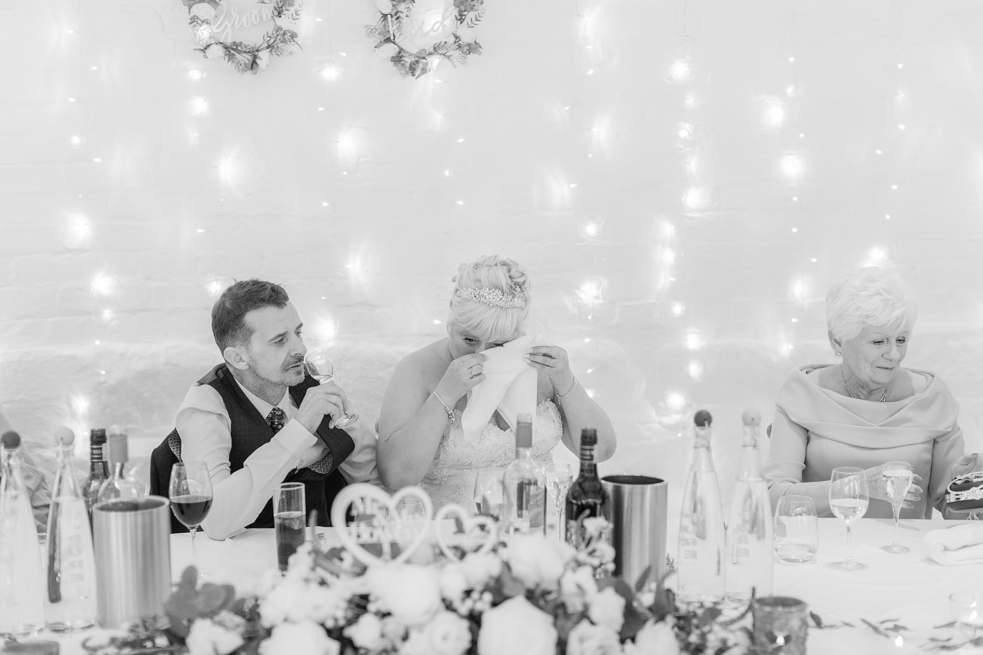 photo shows a bride crying with a napkin to her eyes during the speeches at her wedding, her husband is sat next to her and the other side is her mum
