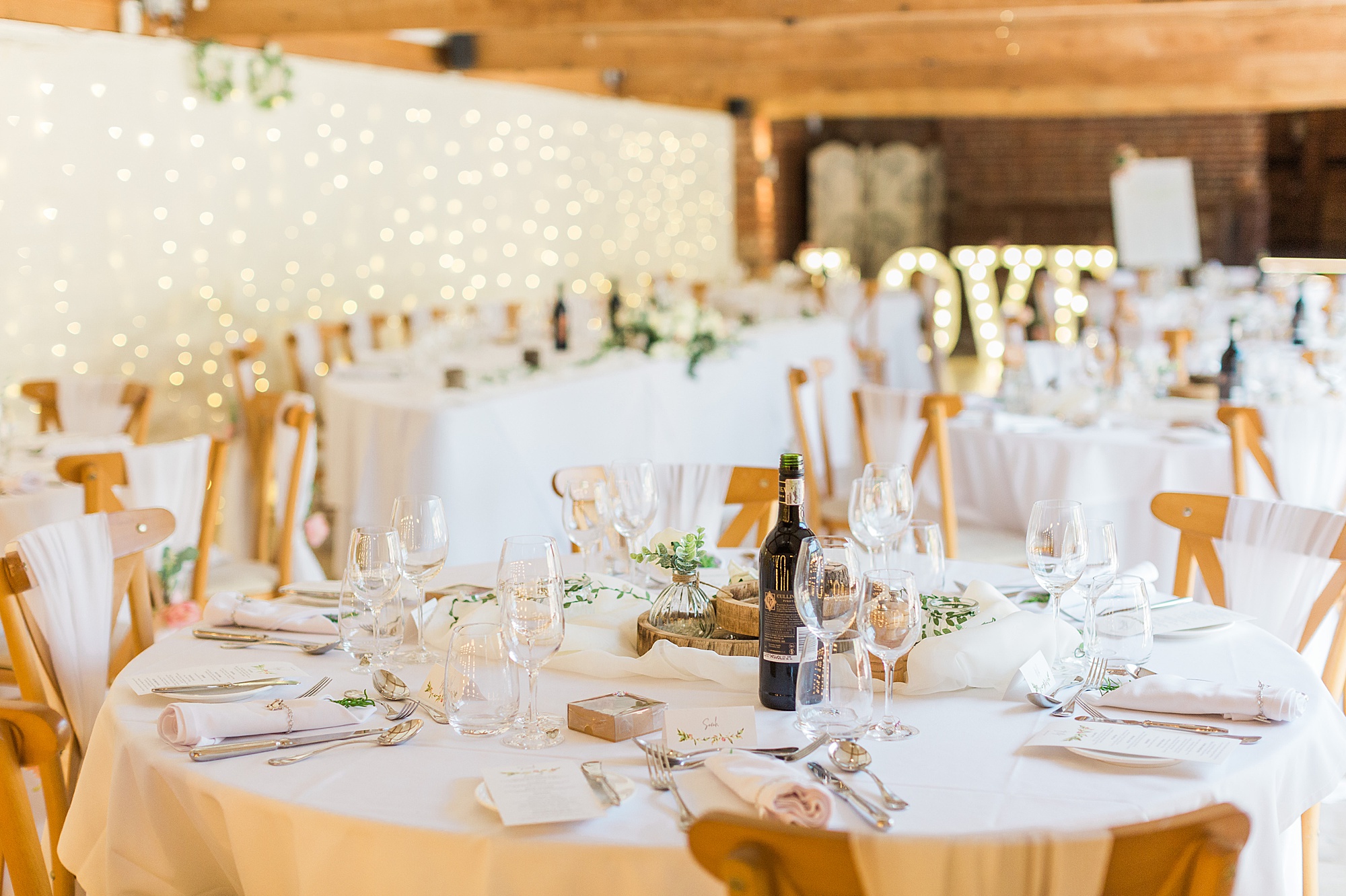 photo of a wedding reception styled beautifully in the barley barn at curradine barns image shows a wall of fairy lights and light up love letters behind simply decorate tables 