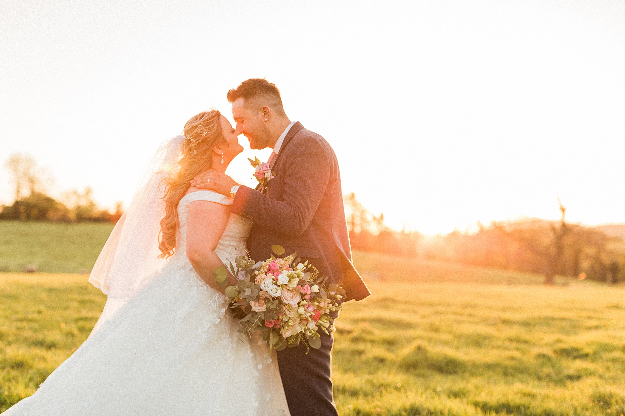 photo of a bride and groom with their noses just touching about to kiss. the groom's left hand is on the bride's shoulder. They are stood in a field with the setting sun behind them 