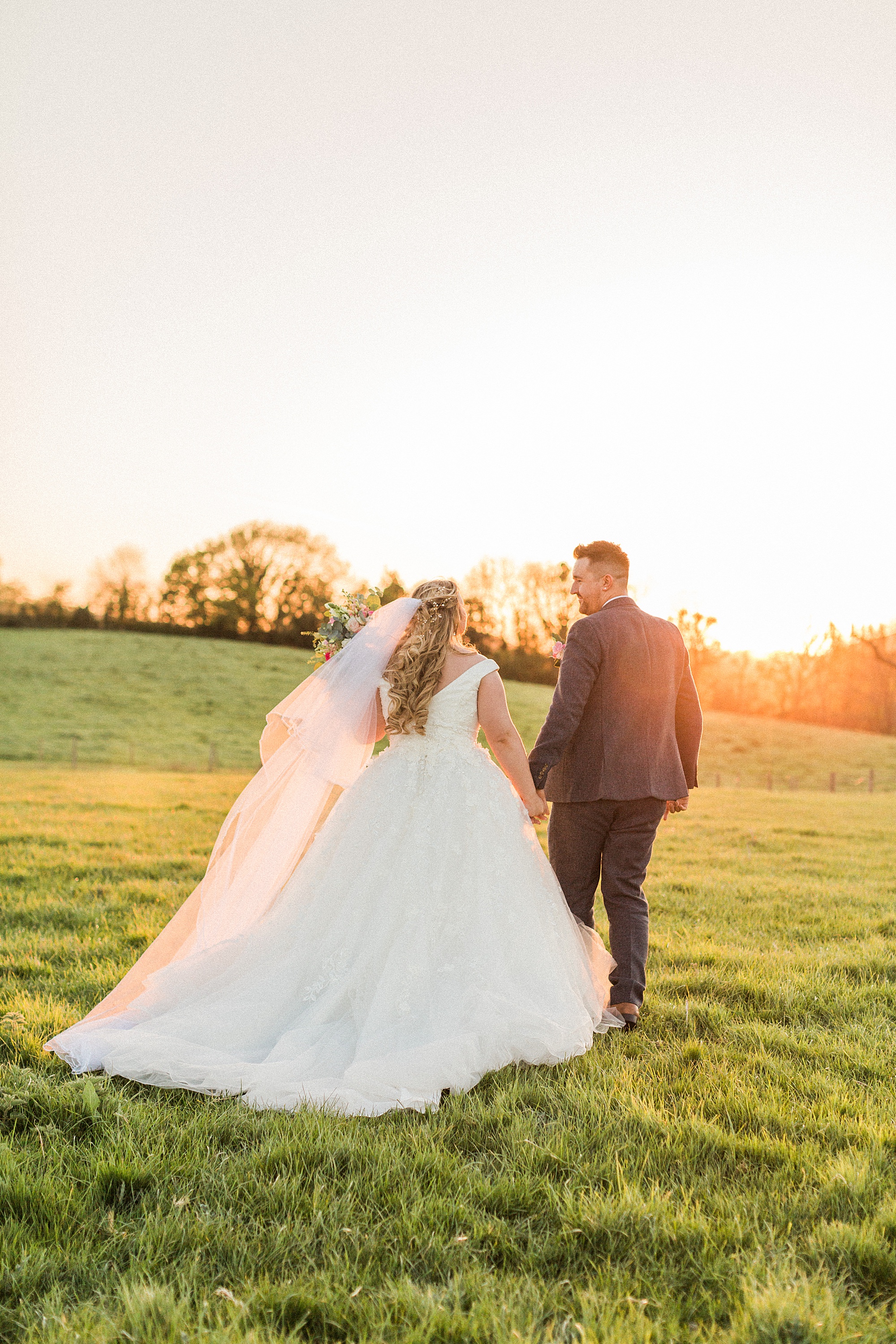 Image shows a bride and groom on their wedding day walking together holding hands in a field during sunset, looking and smiling towards each other. They're walking away from the camera