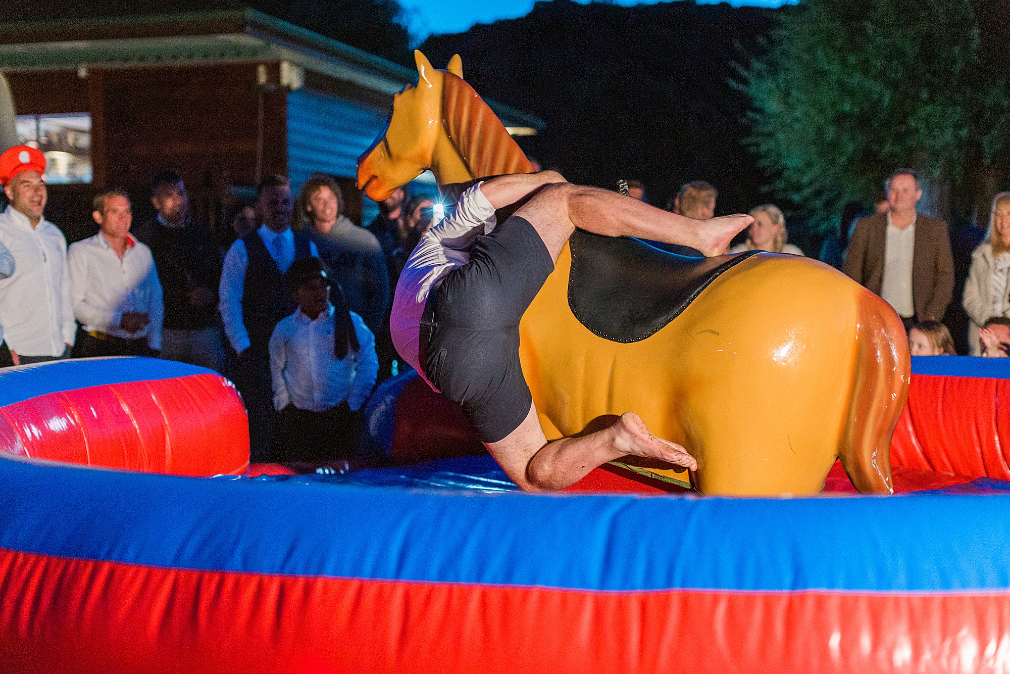 photo of a wedding guest riding a mechanical rodeo pony outside at a wedding reception with guests watching, the groom is falling from the horse and mid way to landing with his bottom and legs facing towards the camera 