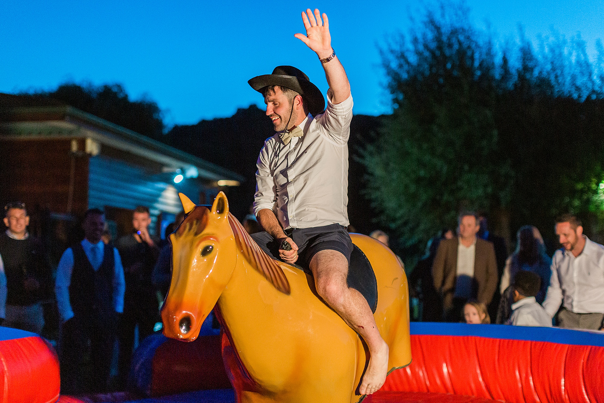 photo of a groom riding a mechanical rodeo pony outside at a wedding reception with guests watching. he is wearing a cowboy hat and has one hand waving up in the air 