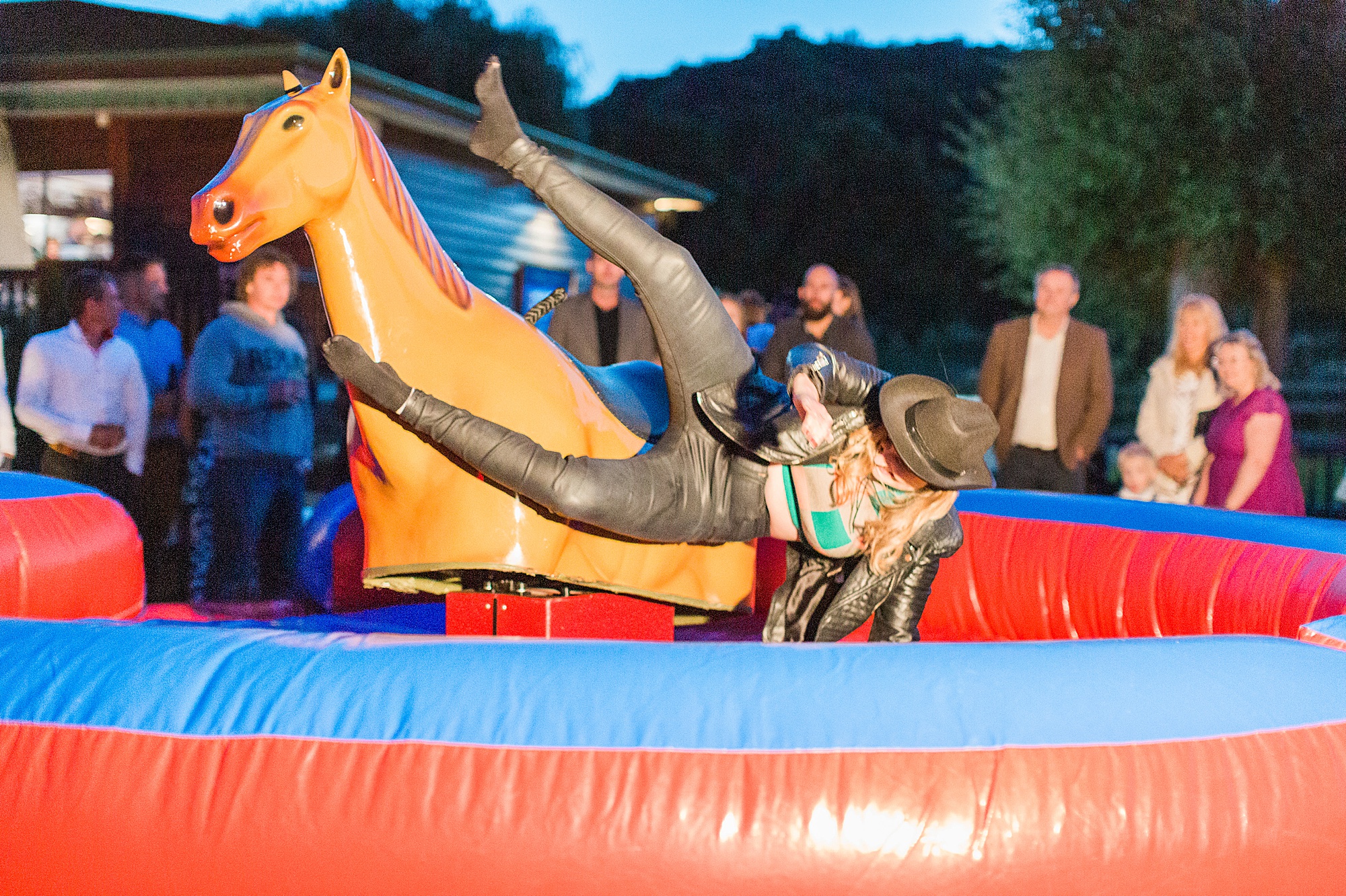 photo of a wedding guest riding a mechanical rodeo pony outside at a wedding reception with guests watching. The guest is almost horizontal to the horse as she falls off 