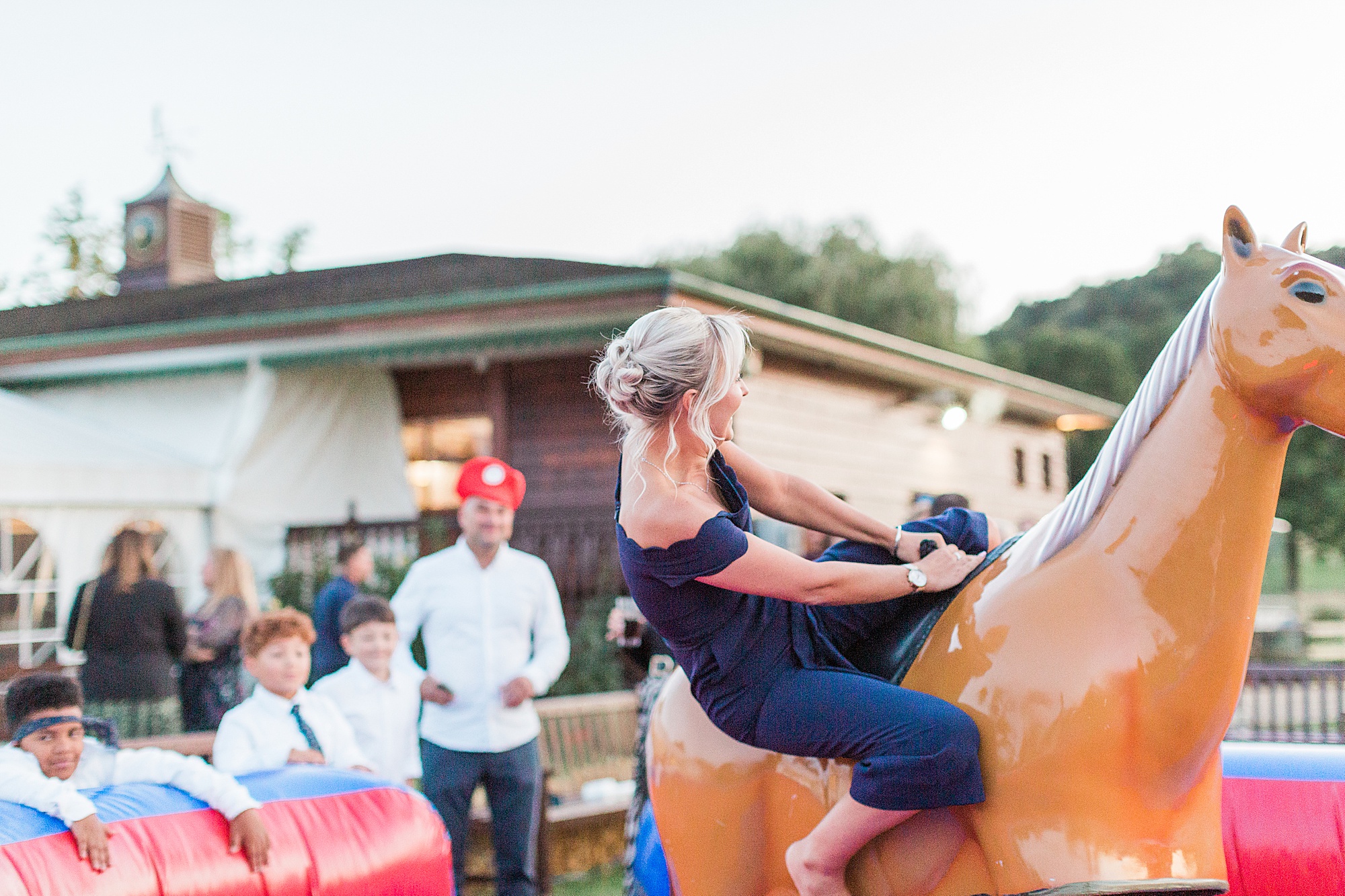 photo of a wedding guest riding a mechanical rodeo pony outside at a wedding reception with guests watching - the female guest is clinging on and half walling from the pony