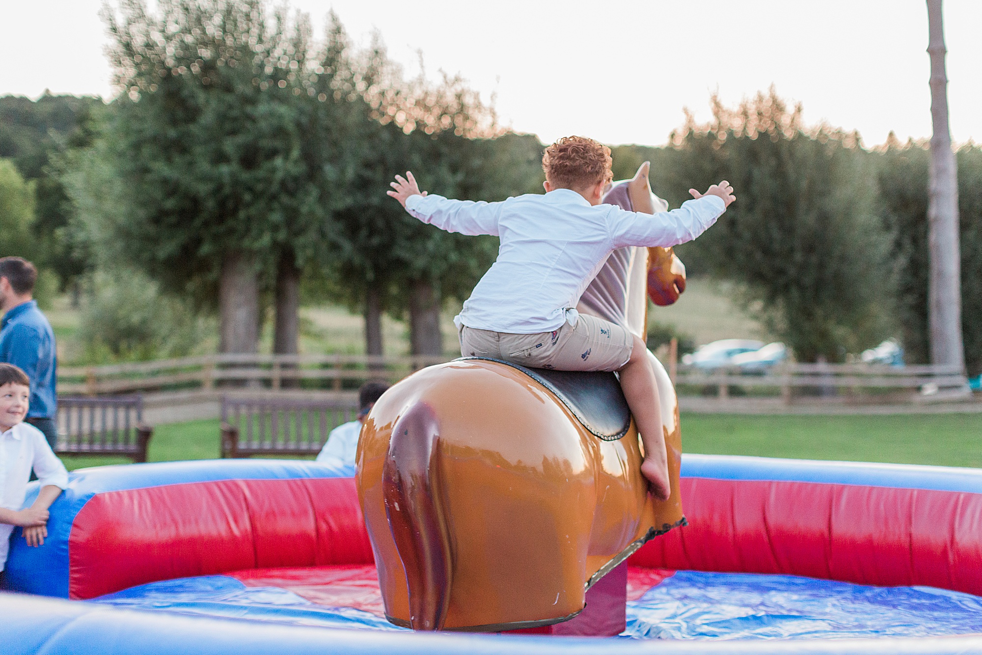 photo of a young wedding guest riding a mechanical rodeo pony outside at a wedding reception with guests watching. the boy has his arms outstretched