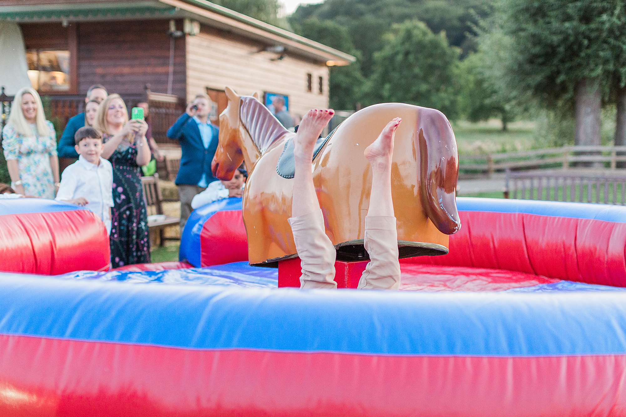 photo of a wedding guest riding a mechanical rodeo pony outside at a wedding reception with guests watching, the person has fallen off the pony and landed with their feet in the air