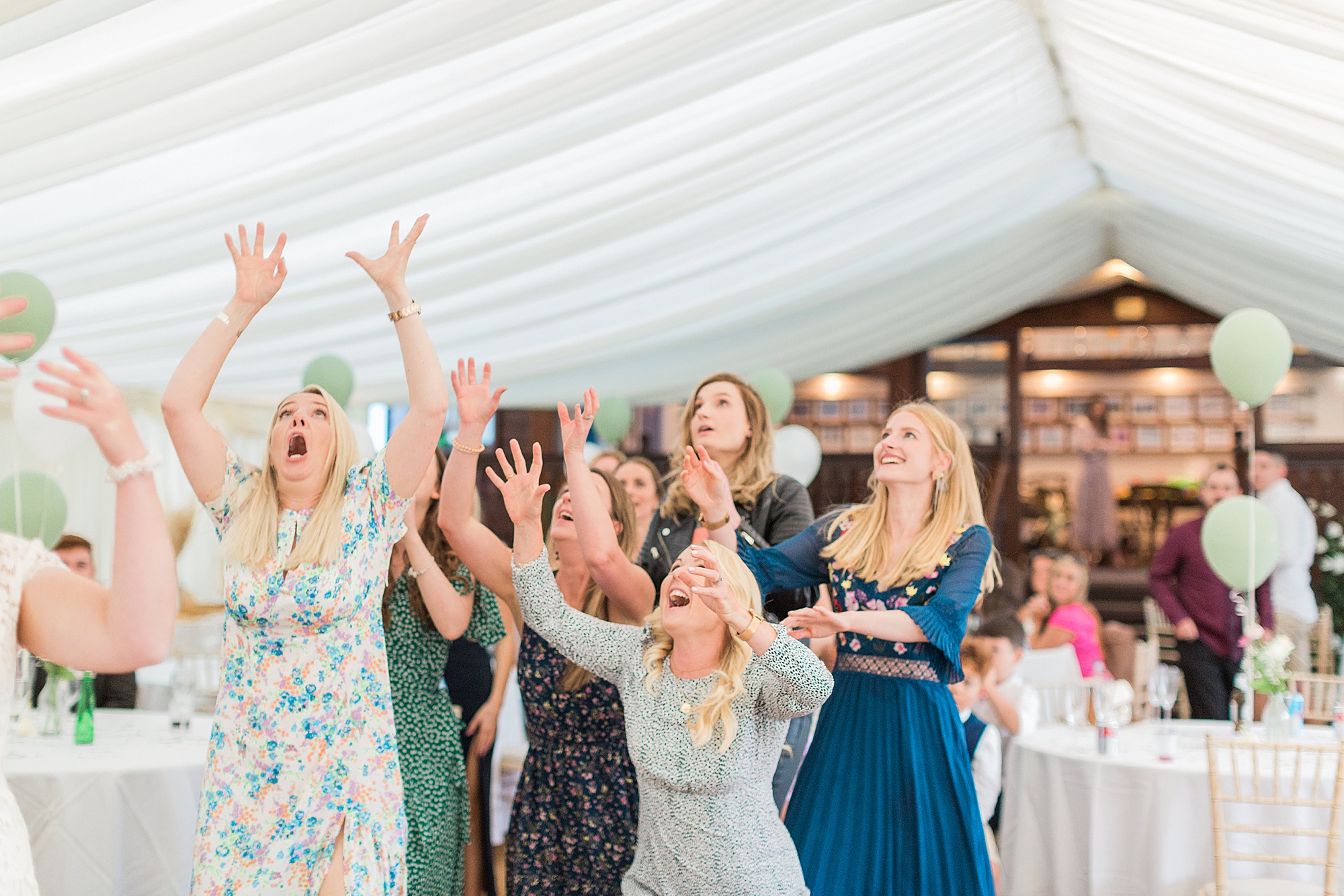 photo shows a group of female wedding guests all rushing with their arms out ready and poised to catch the bride's bouquet that has been thrown