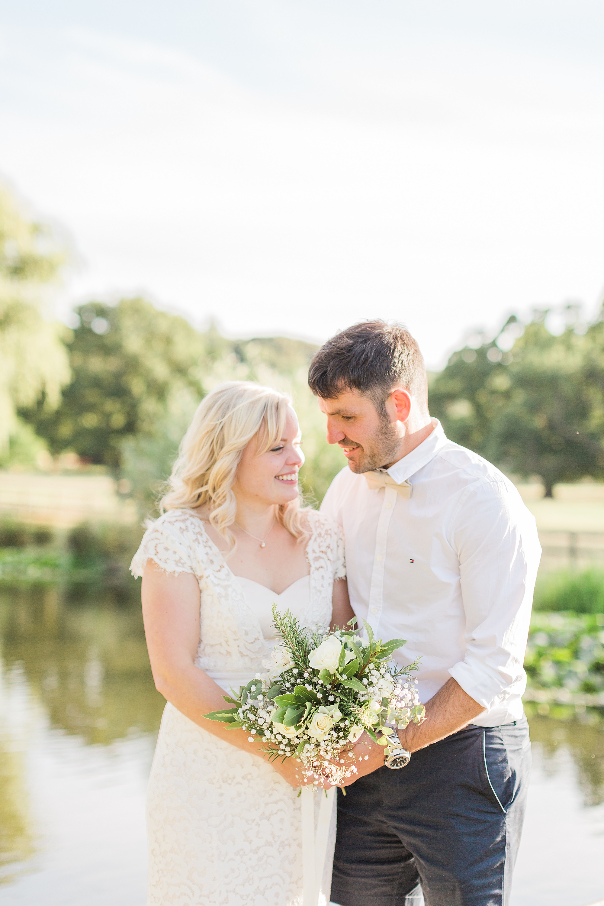 photo of a bride in a lace dress and groom wearing shirts and shorts stood embraced in towards each other holding the bride's bouquet with a lake in the background