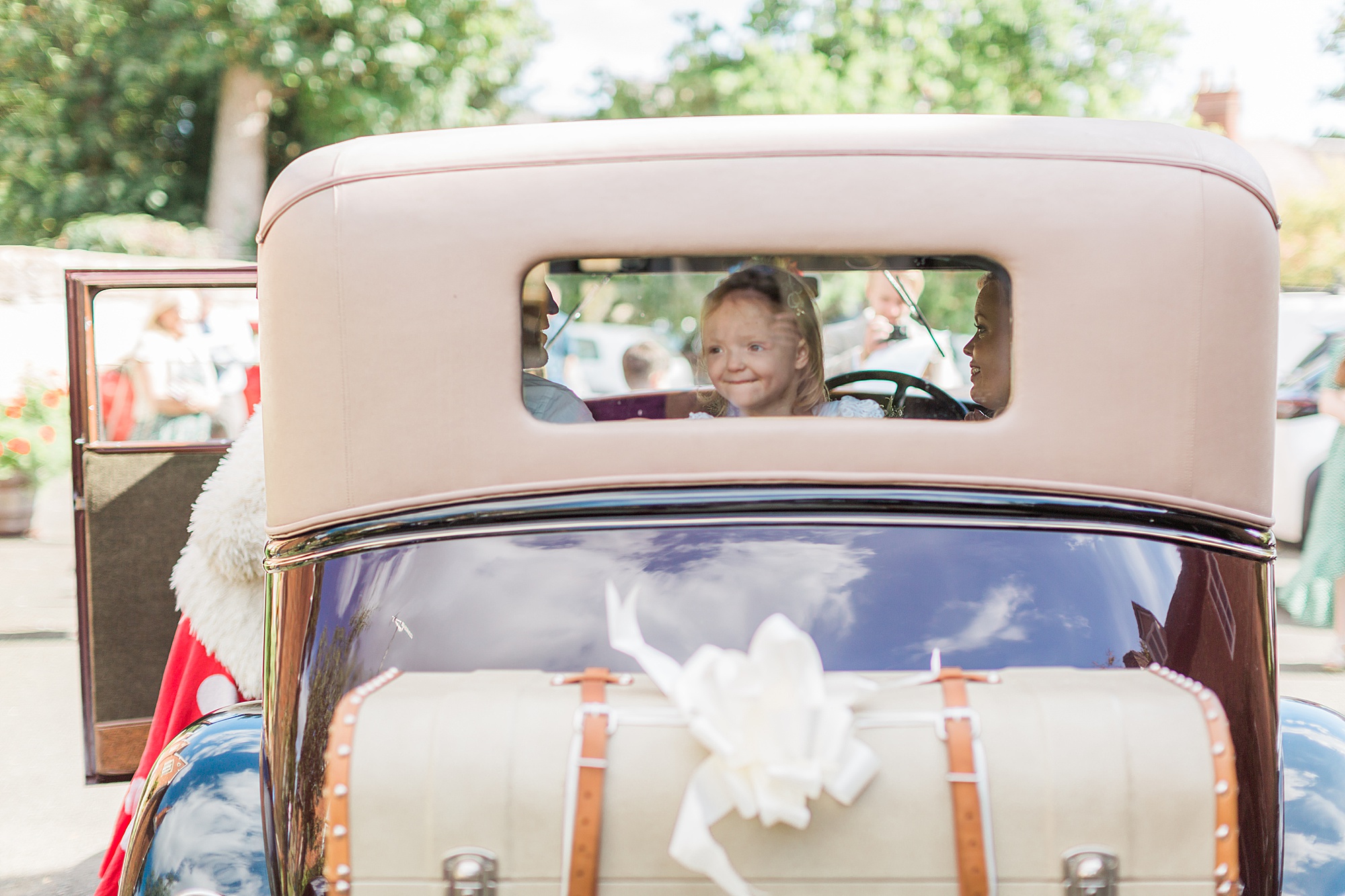 photo of a flower girl looking through the back window of a vintage car leaving a wedding in the cotswolds