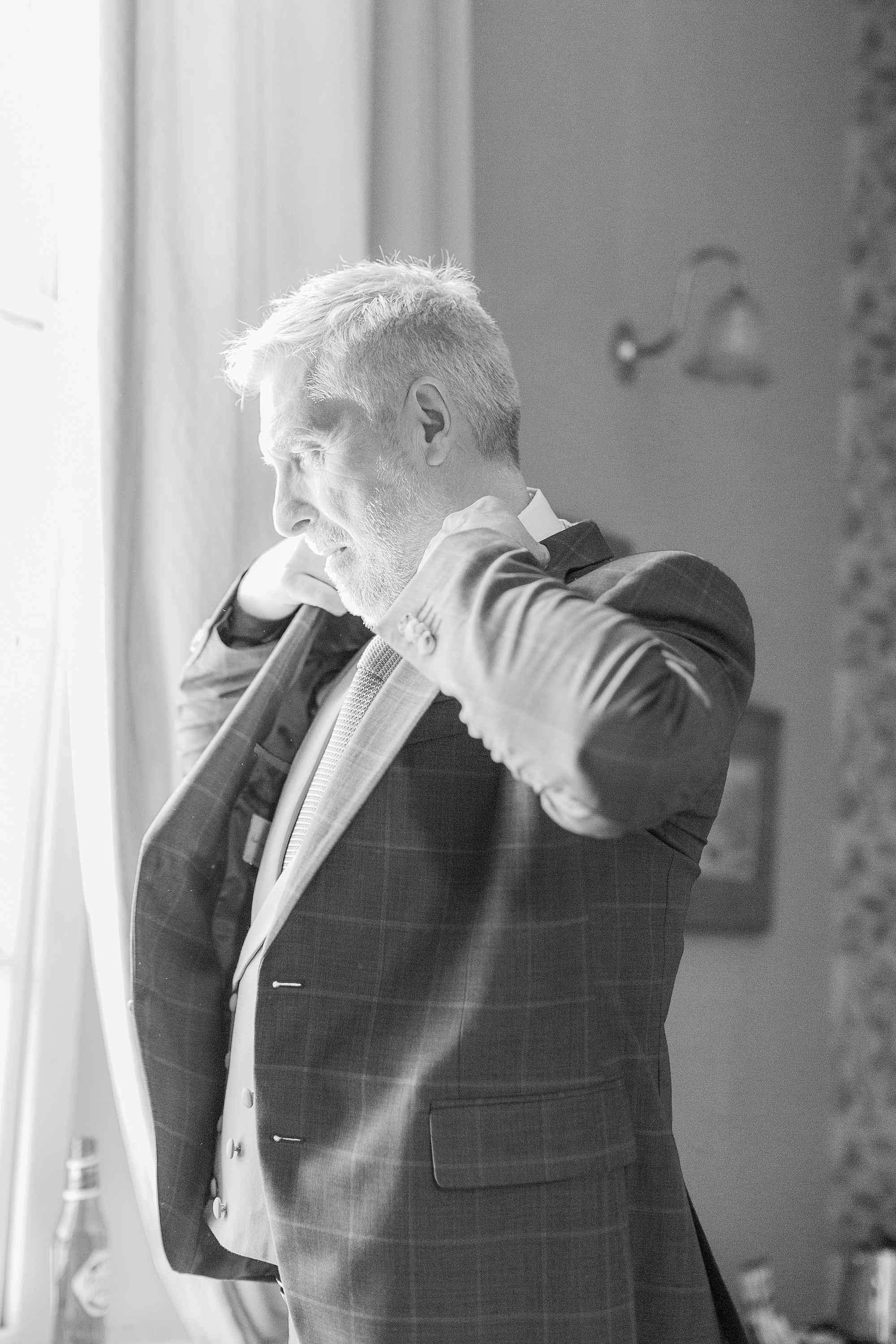 photo of a groom looking out of the window at groton manor on the morning of his wedding, the groom is putting on his suit jacket 