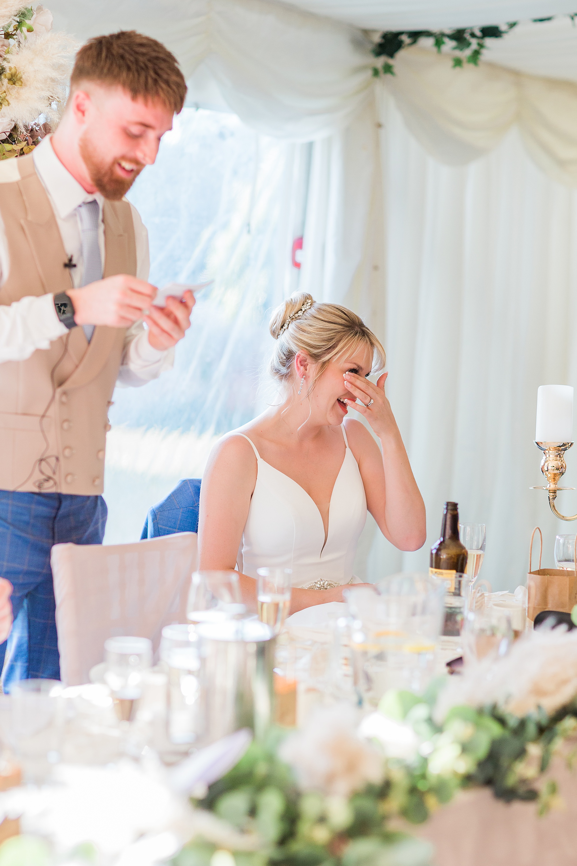 photo of a best man speaking during the wedding's speeches and the bride holding her hand to her face covering her face slightly and laughing 