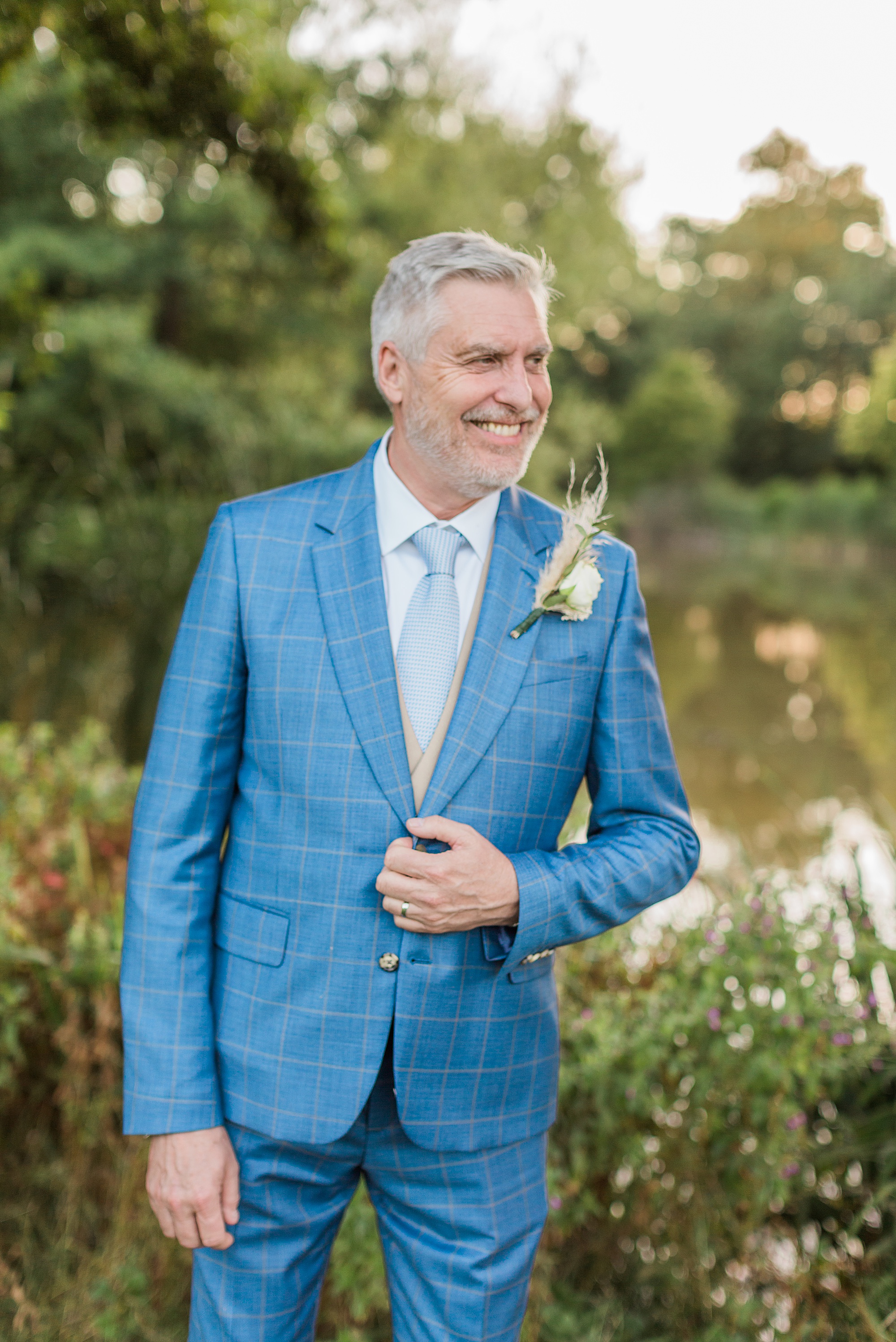 photo of a groom looking towards his wife (not pictured), groom is wearing a blue suit and holding his jacket with a smile on his face. He's stood in front of a lake 