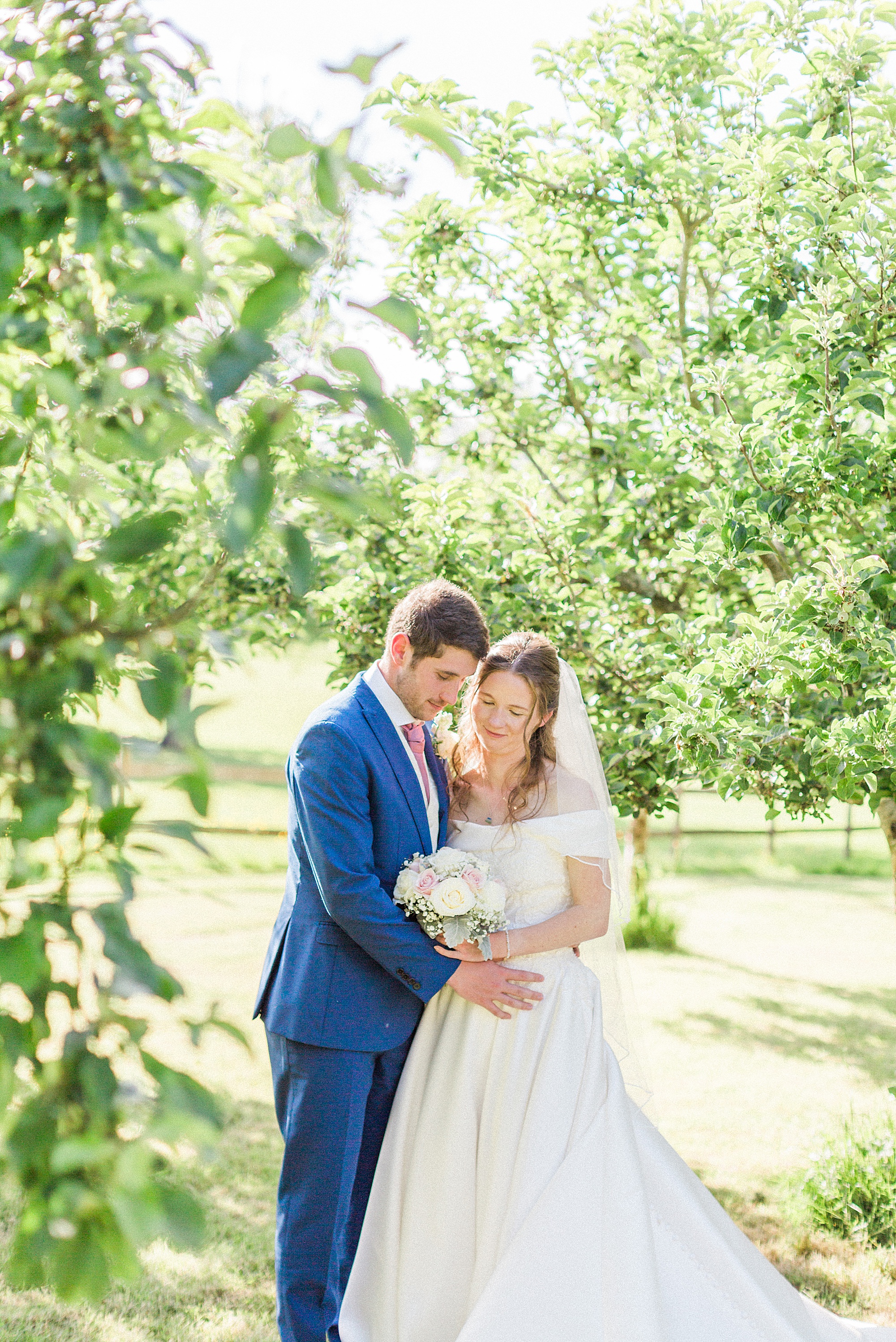 bride and groom being photographed in an orchard, couple are embraced and cuddled in towards each other 