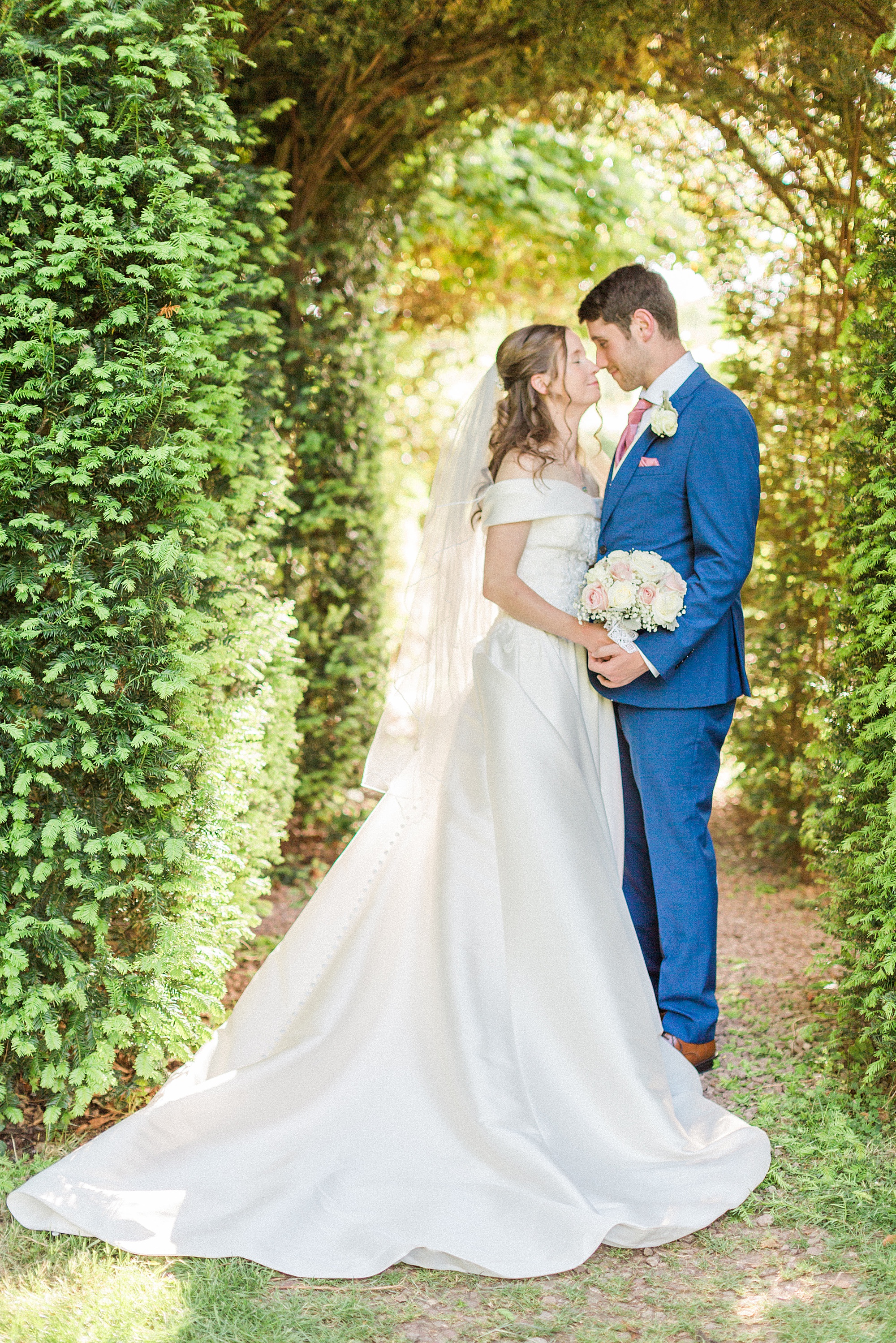 photo of a bride and groom facing in towards each other connected with their faces almost touching and bodies/hands together. They are lit from behind with golden sun and the archway is evergreen greenery