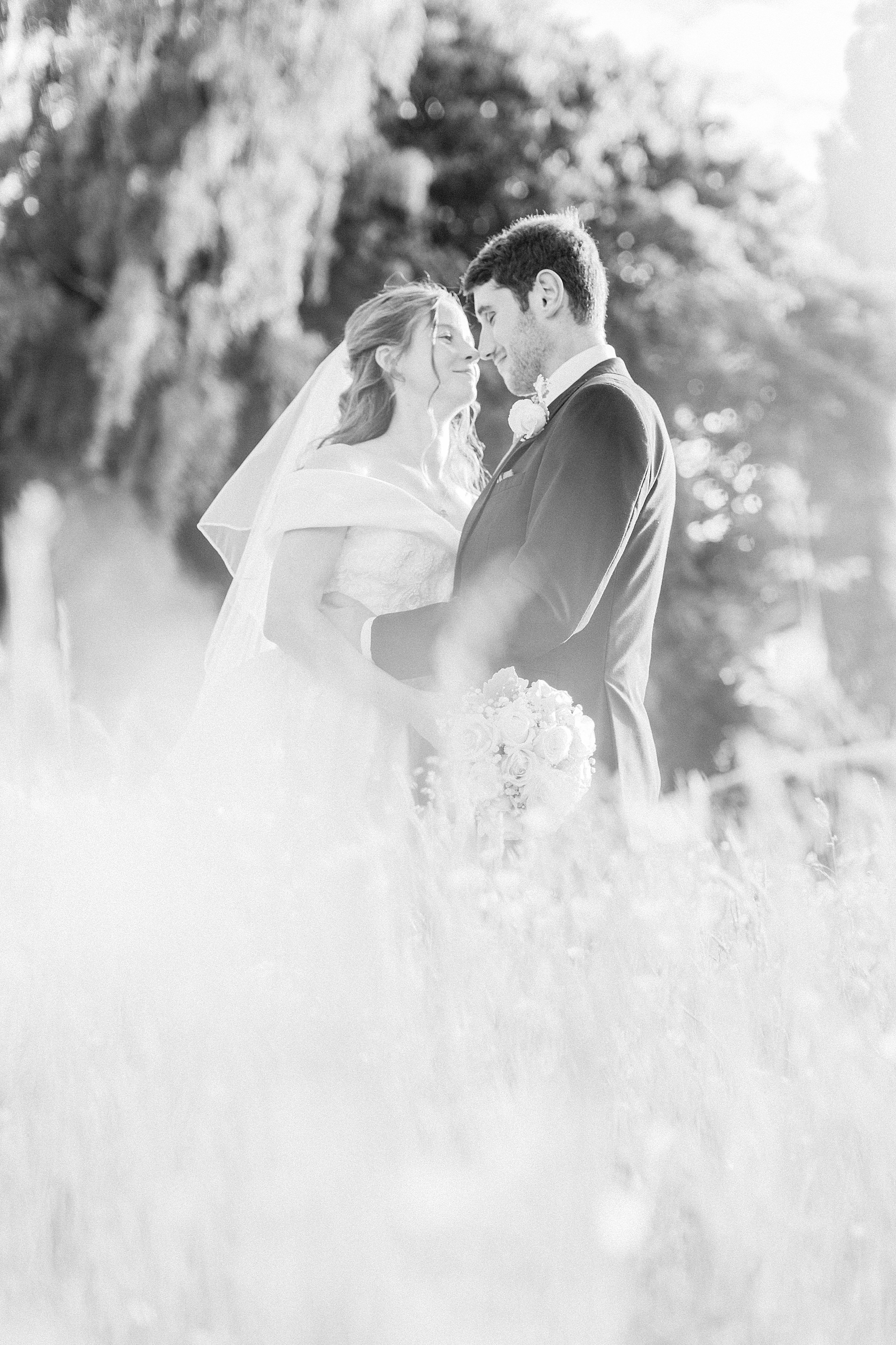 black and white photo of a bride and groom stood in very long grass in a field with the noses touching and facing in towards each other smiling whilst embraced