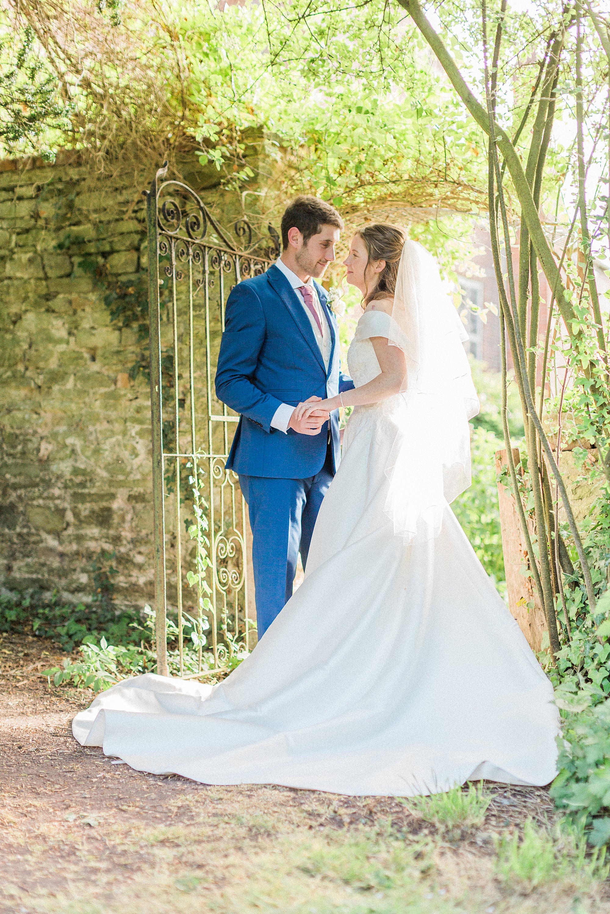 photo of a bride and groom facing towards each other holding hands in a gateway with wall and trees surrounding them