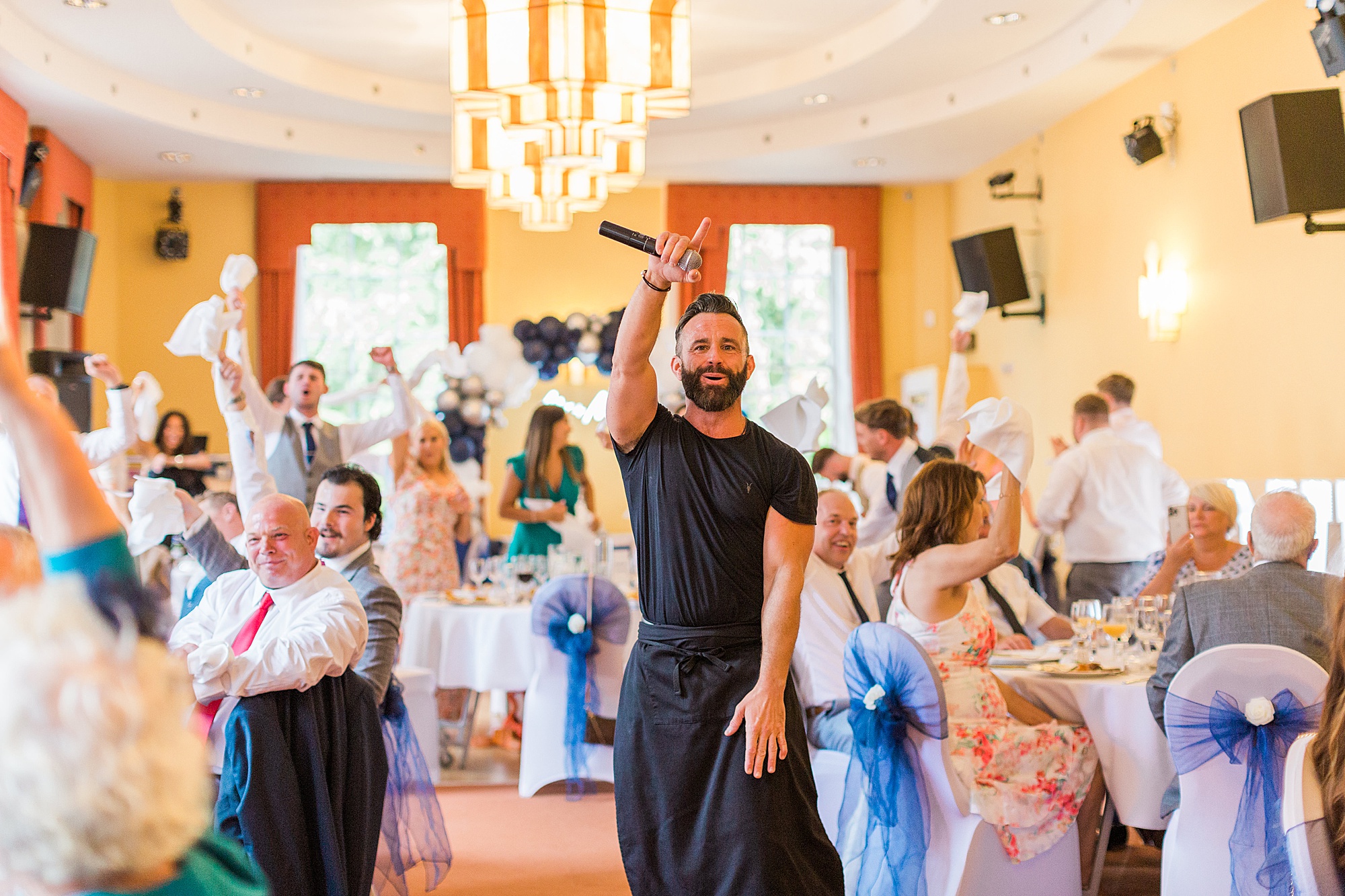  singer waiter with his hand up in the air performing to wedding guests with guests in the background singing and cheering 