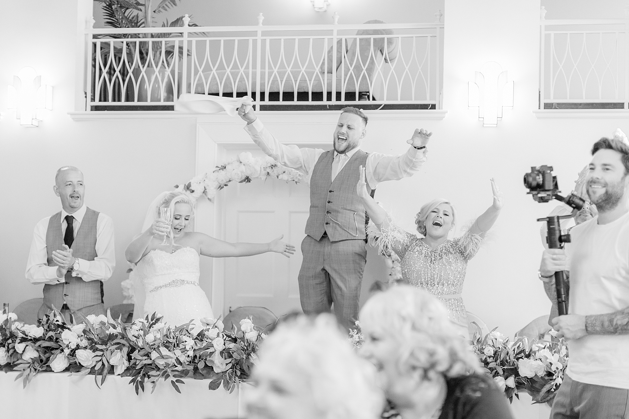 groom at a wedding standing on a chair with his hands in the air cheering and singing to singing waiters, bride and mum and groom to each side of him cheering also 