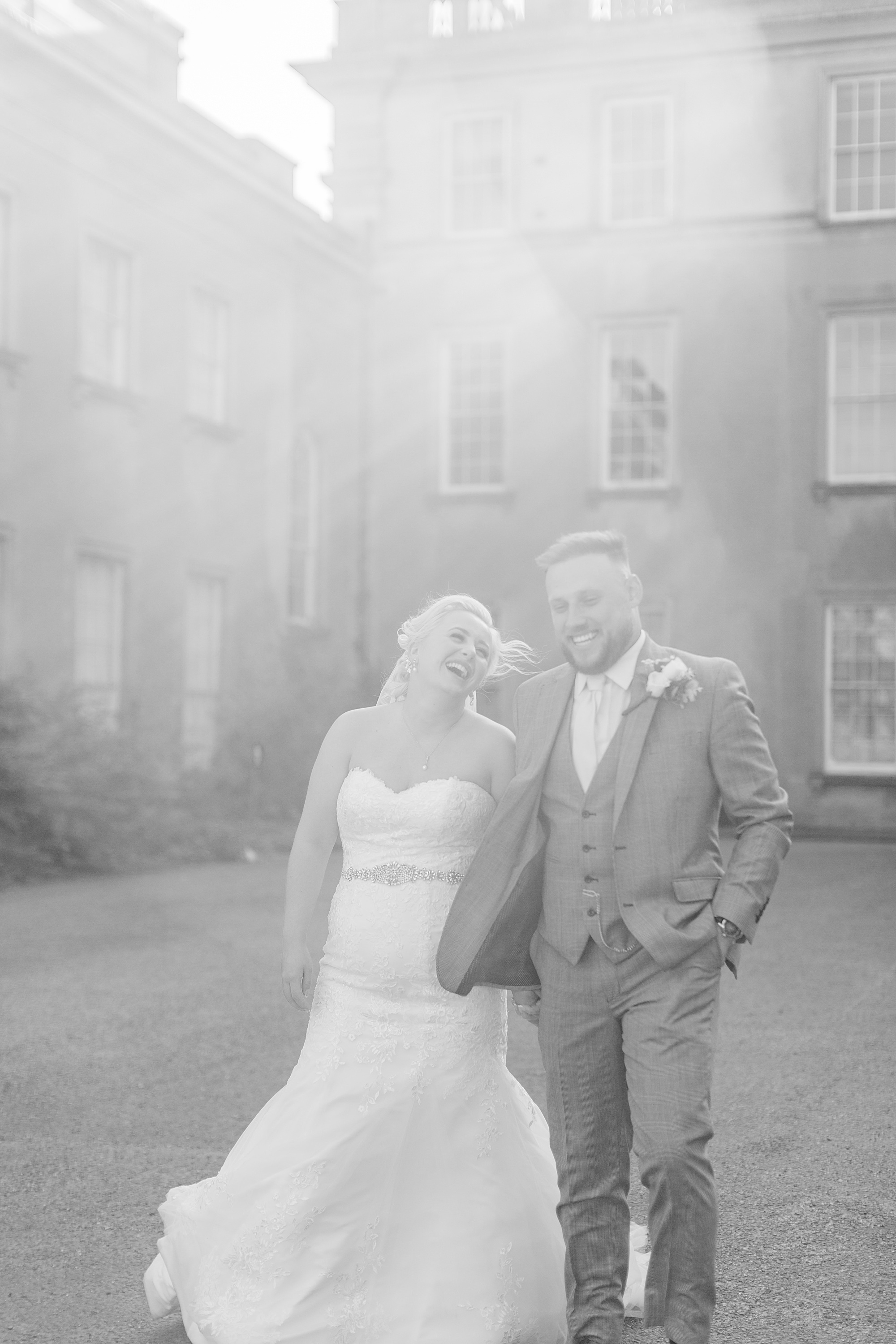 bride and groom walking with himley hall behind them and the sunset and soft lighting and glare from the sun in the image behind them too