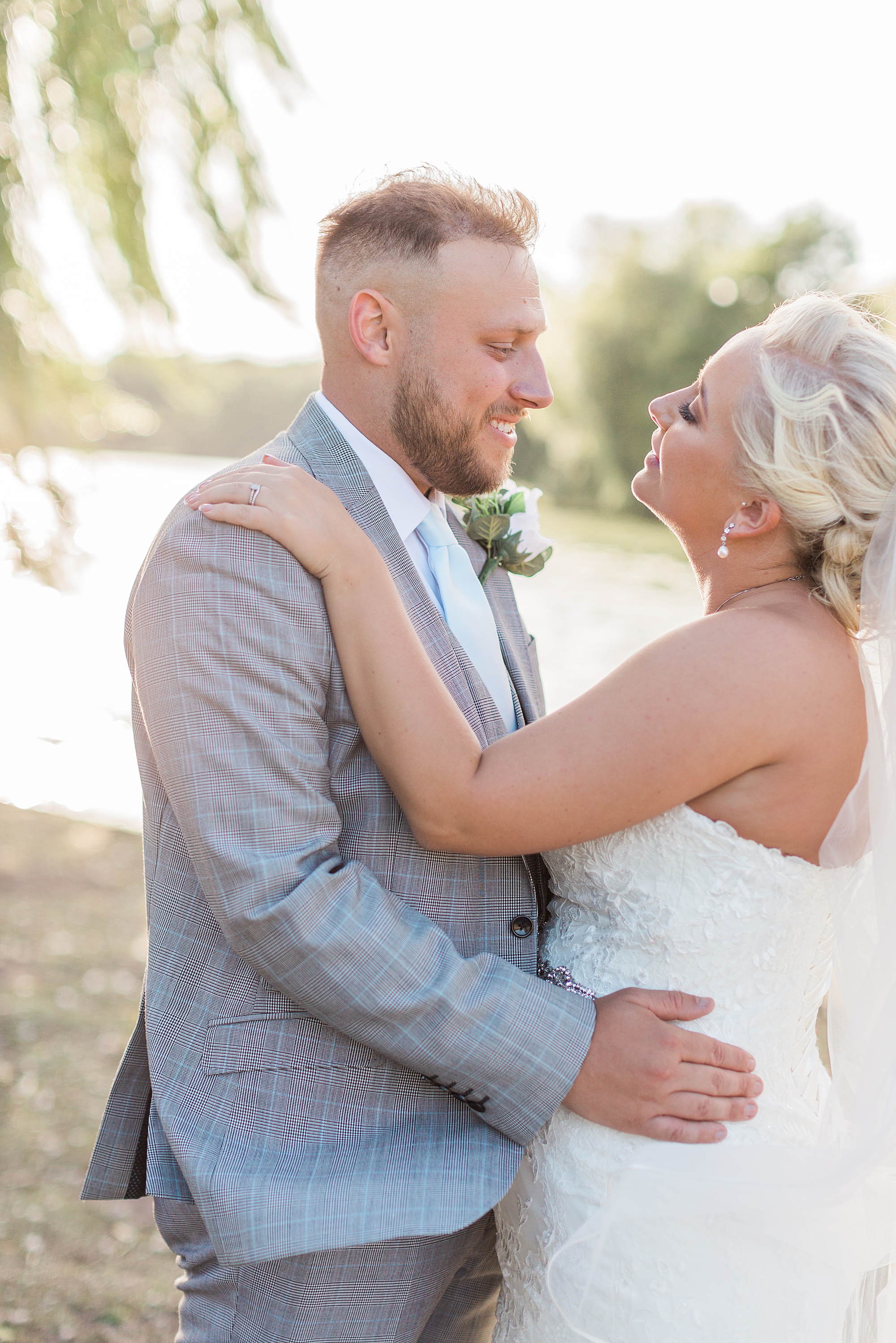 Photo of a bride and groom embraced and looking towards each other stood under a tree and in front of a lake with the sun shining behind them