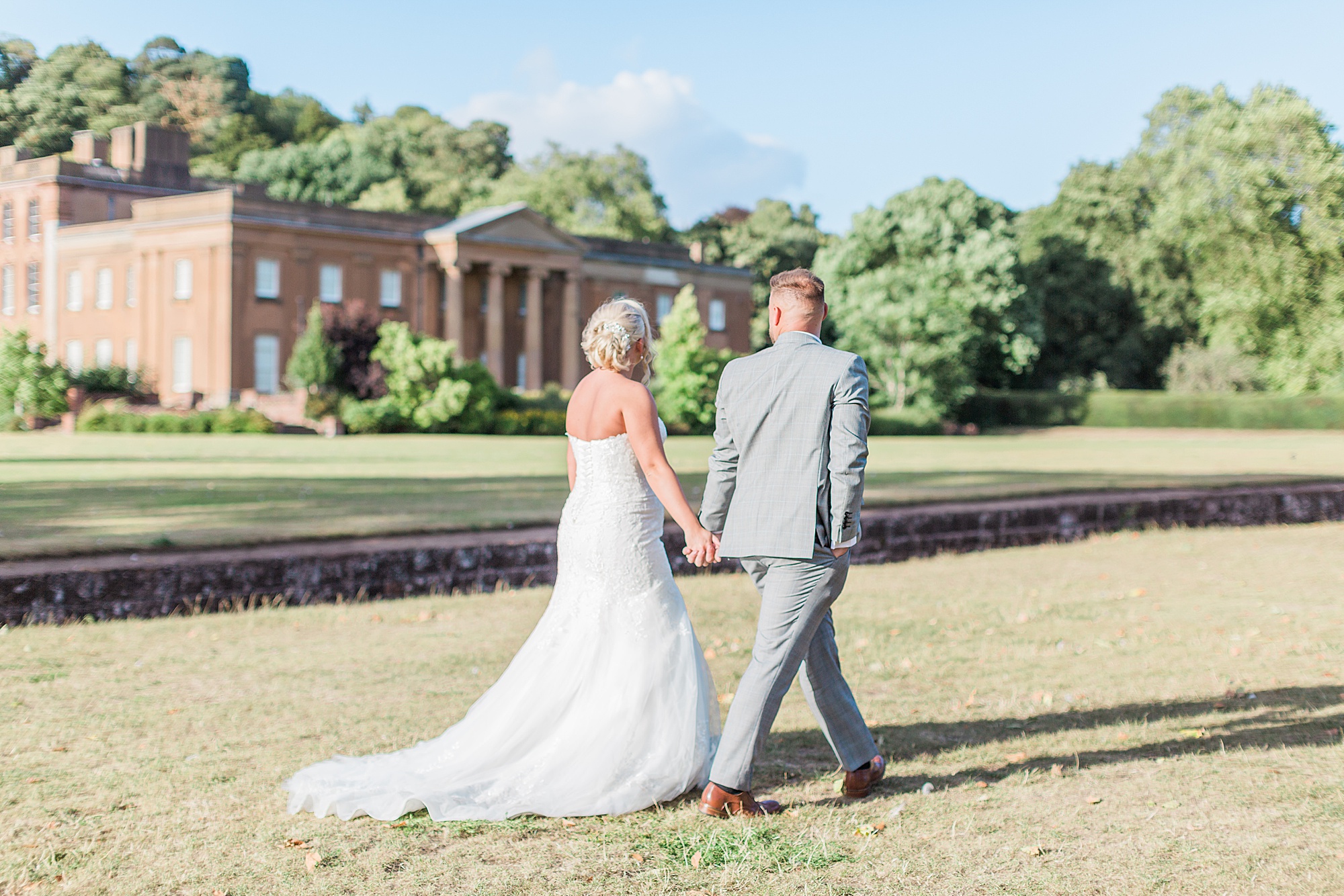 bride and groom walking hand in hand along the lawn in front of himley hall. Couple are talking and looking towards the venue in the evening sun 