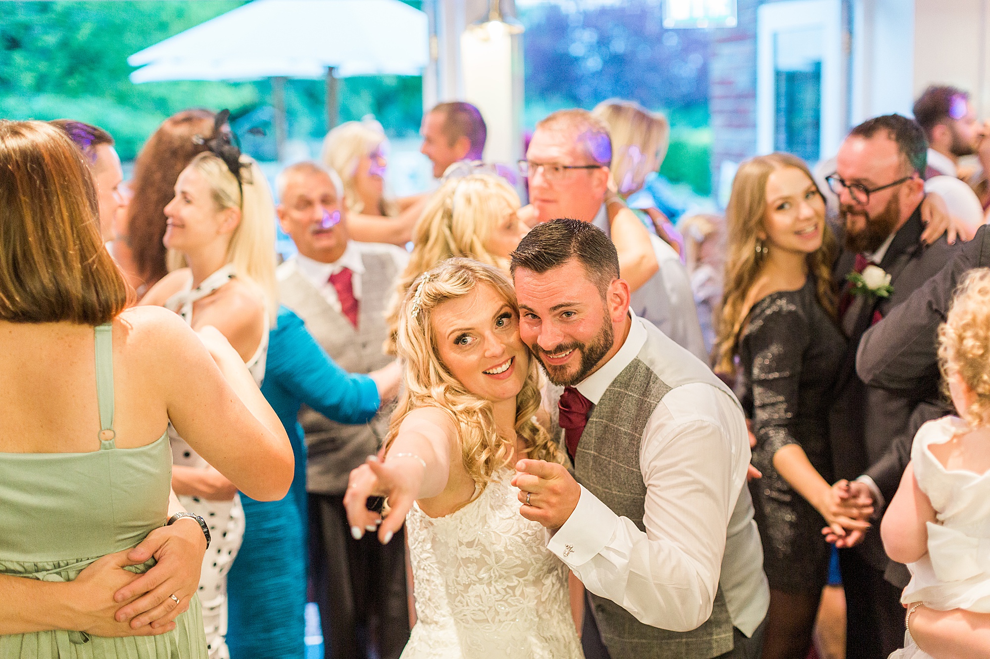 image shows guests dancing and singing at a wedding reception in the evening including a bride and groom pointing to another guest to come and dance