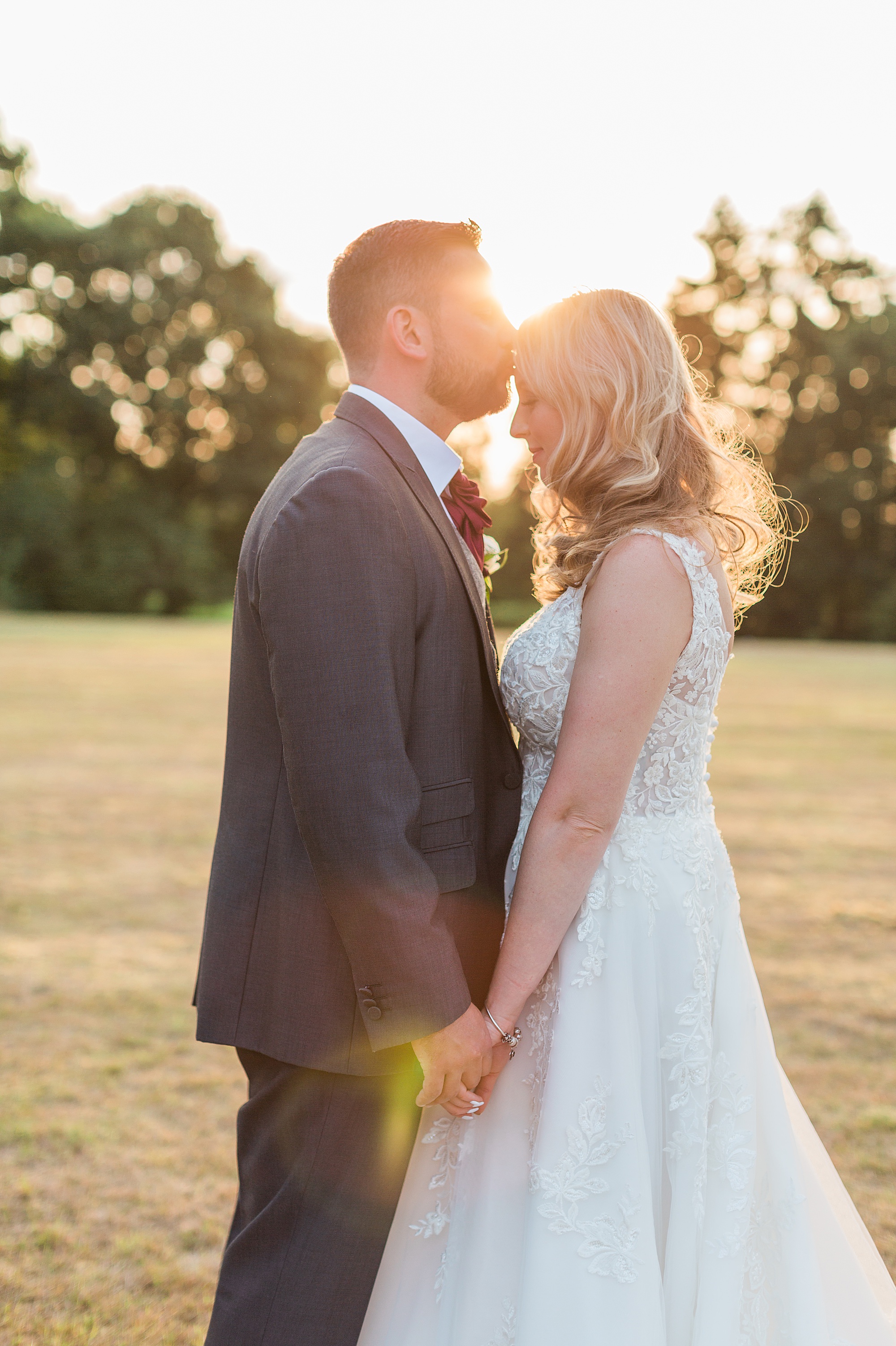 photo of a bride and groom stood facing towards each other and their bodies connected, holding hands with the setting sun behind them. the groom is kissing his wife on the forehead