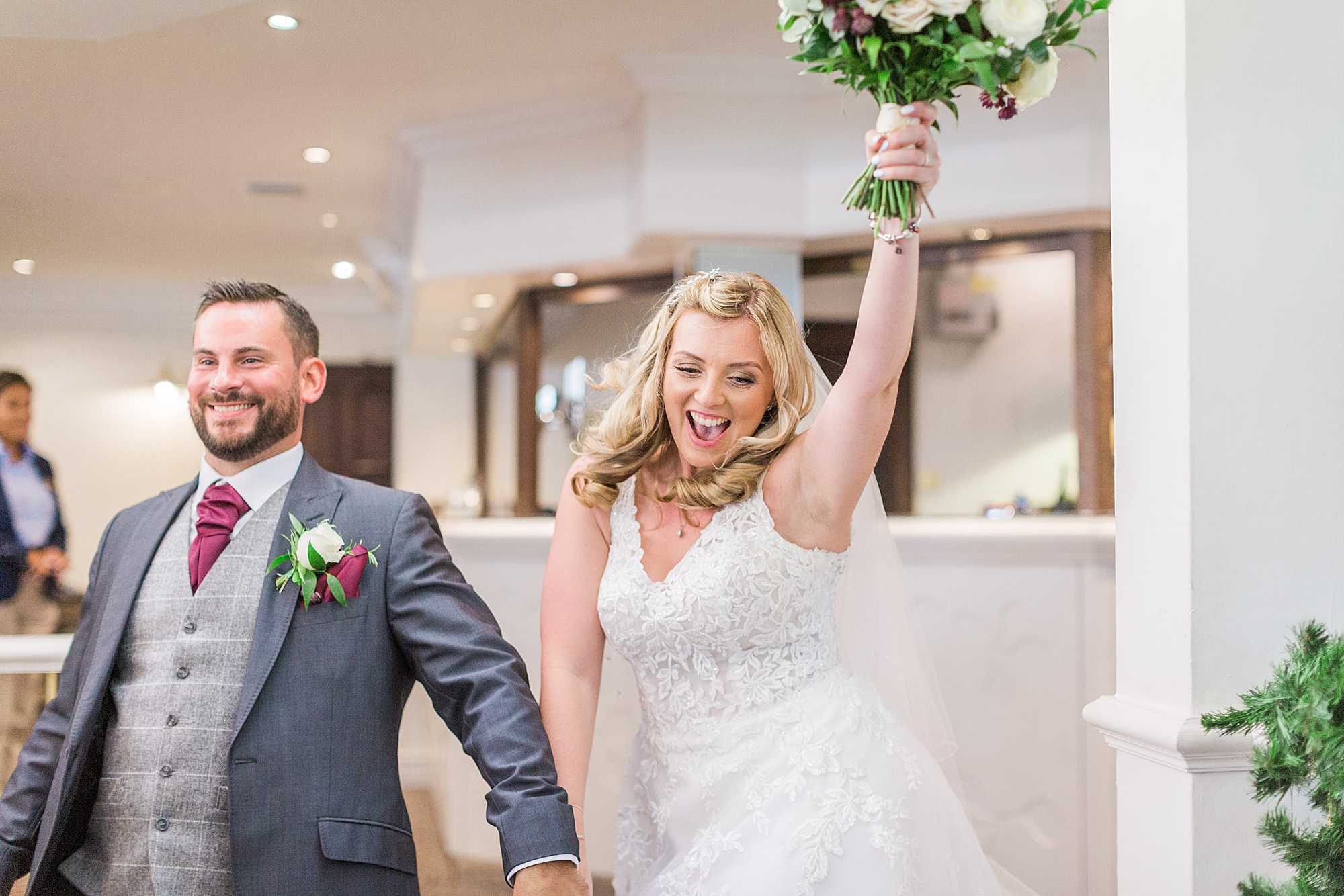 image shows a bride and groom entering their wedding reception holding hands, the bride is punching her bouquet up into the air 